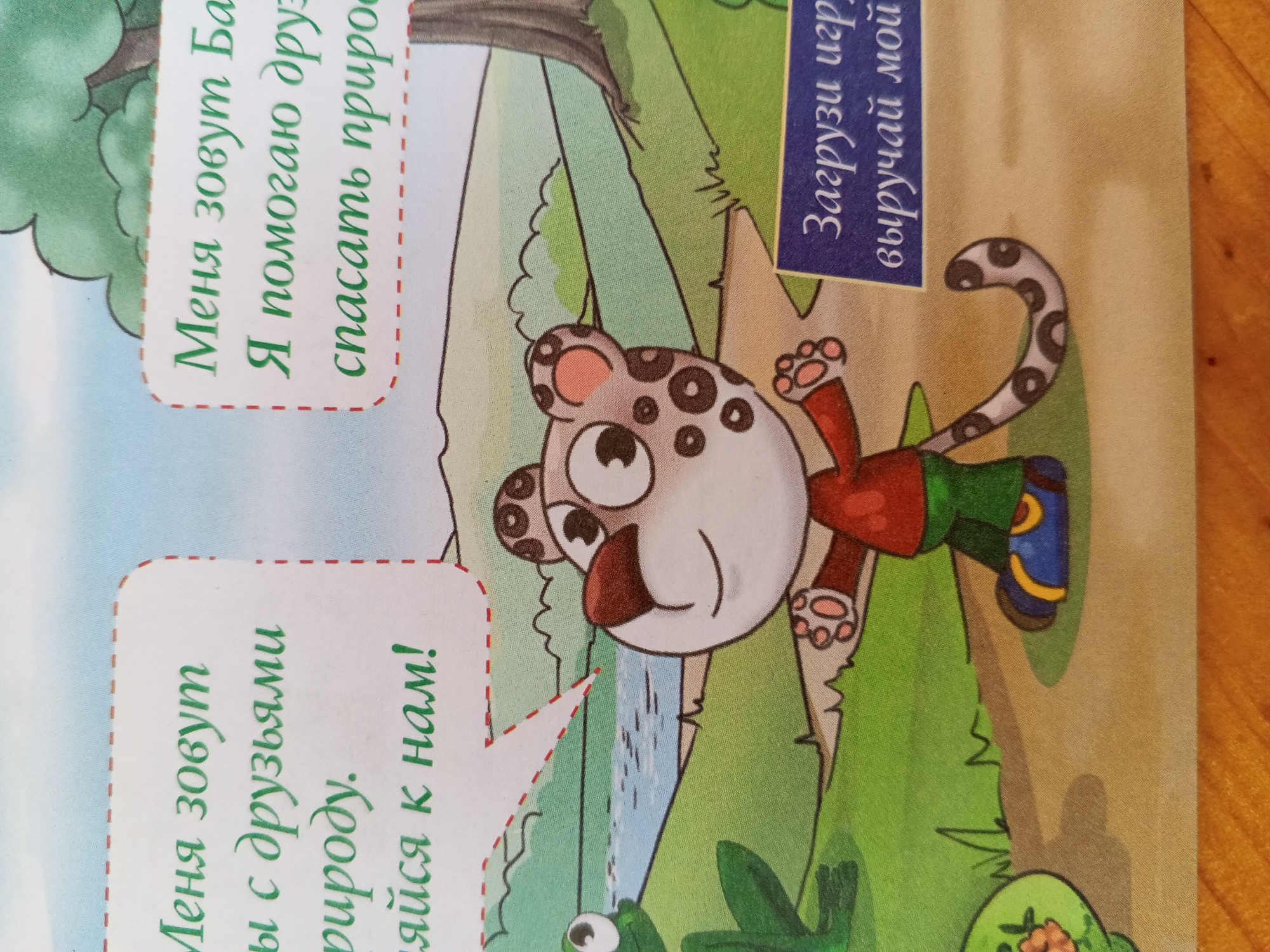 The image depicts an illustration from a children's book. On the left side, a white speech bubble with a red dashed border contains Russian text. The text reads: "Меня зовут Ба Я помогаю дру спасать приро". To the right of the speech bubble is a cartoon animal, possibly a leopard or cheetah cub, with spots, large eyes, and wearing a red shirt and blue pants with yellow accents. The animal is in a dynamic pose, appearing to be jumping or climbing. Behind the animal is a green, natural landscape with trees and a body of water. In the upper right corner, there is a blue rectangular label with Russian text that reads: "Загрузи игр выручай мо". The image is a photograph of a page from a book, with a wooden surface visible on the right edge. The orientation of the text suggests the image has been rotated. There is no visible indication of the city of Chistopol, Russia.