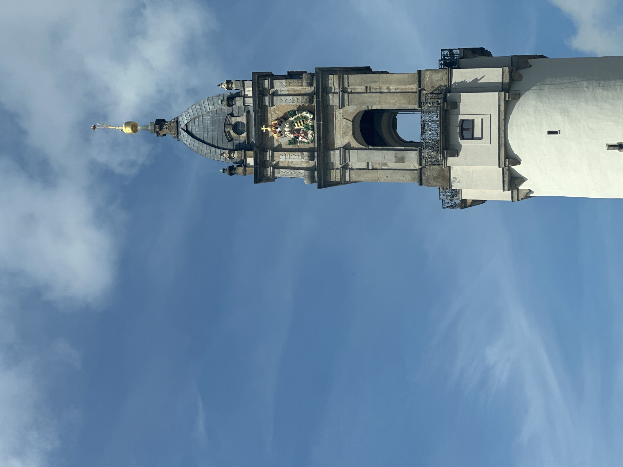 The image shows a close-up view of the Reichenturm, a historic tower located in Bautzen, Germany. The tower is tall and slender, with a white cylindrical base that tapers as it rises. Above the base, there are several architectural features including a small rectangular window and a balcony with ornate metal railings. Further up the tower, a section of stone construction is visible, featuring an arched opening and decorative stonework. At the very top of the tower is a pointed roof with a golden spire. The sky is blue with scattered white clouds. The scene is captured from a low angle, emphasizing the height of the tower against the sky.