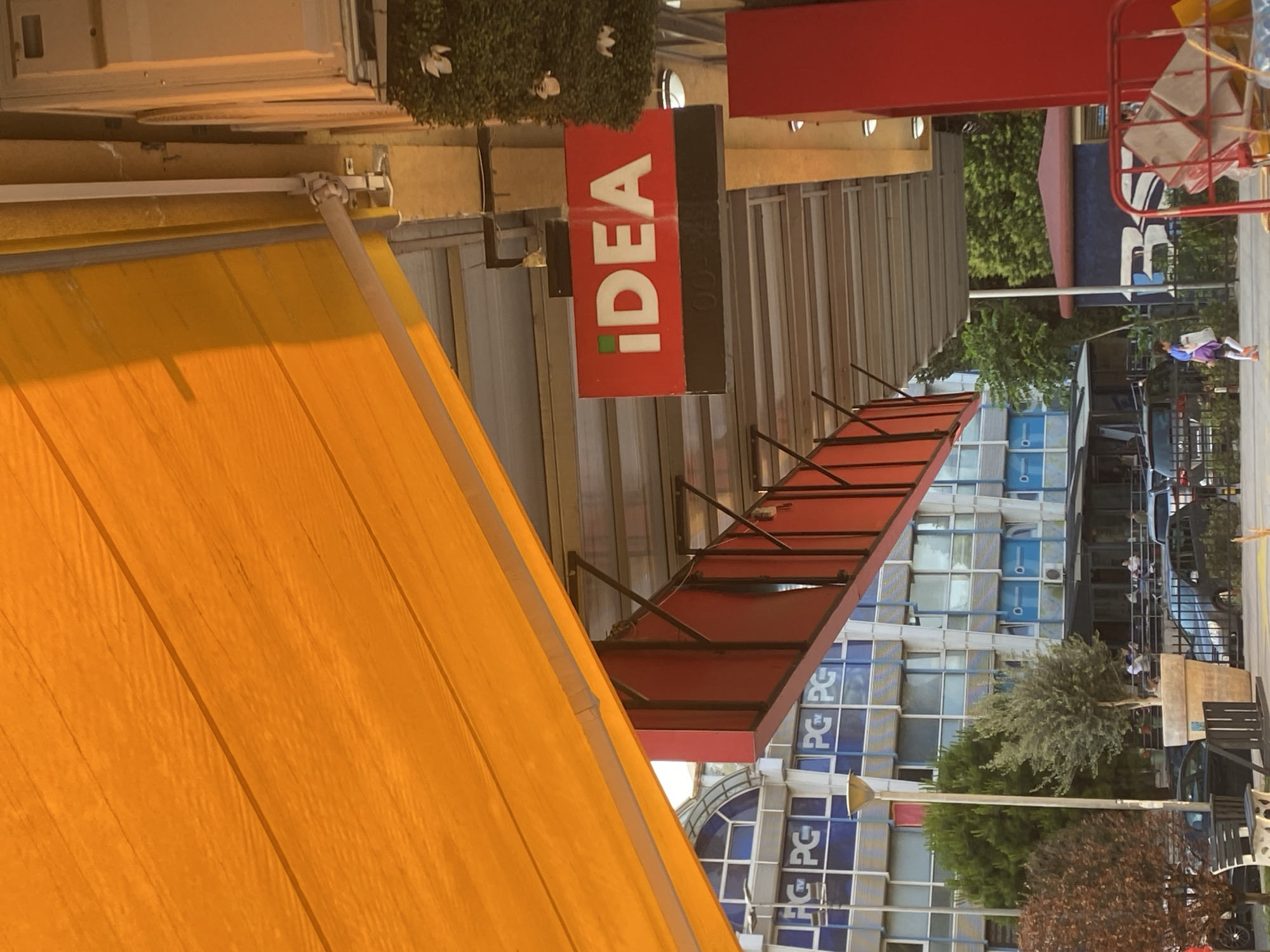 The image shows a street scene in Podgorica, Montenegro, with an "IDEA" sign visible. There is a yellow awning in the foreground and a red awning further back. The building in the background has a modern design with blue and white accents. A person in a purple shirt and white pants is walking on the sidewalk. There are cars parked along the street and trees providing shade. The sky is bright.