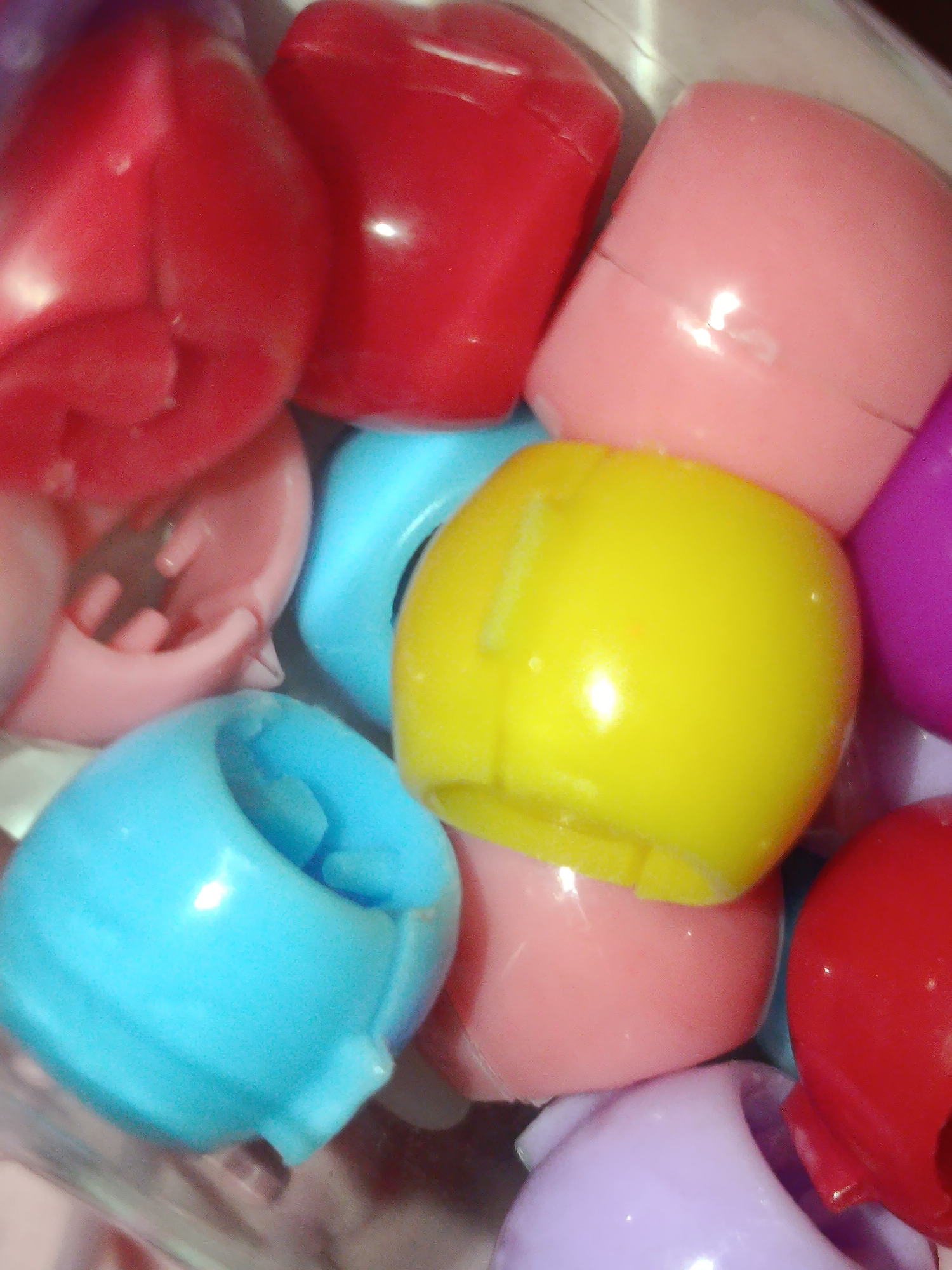 The image shows a close-up, overhead view of a collection of colorful plastic caps, possibly from pens or markers, piled together in a container. The caps are in various shades of red, pink, blue, yellow, and purple. Some are stacked on top of others, and the overall impression is one of a jumbled, vibrant assortment. The lighting is somewhat harsh, creating reflections on the glossy plastic surfaces. The background is out of focus but appears to be a lighter color, possibly the inside of the container. There are no people or any identifiable geographical features present in the image. Therefore, it is impossible to determine the location in Tashkent, Uzbekistan, or any other city.