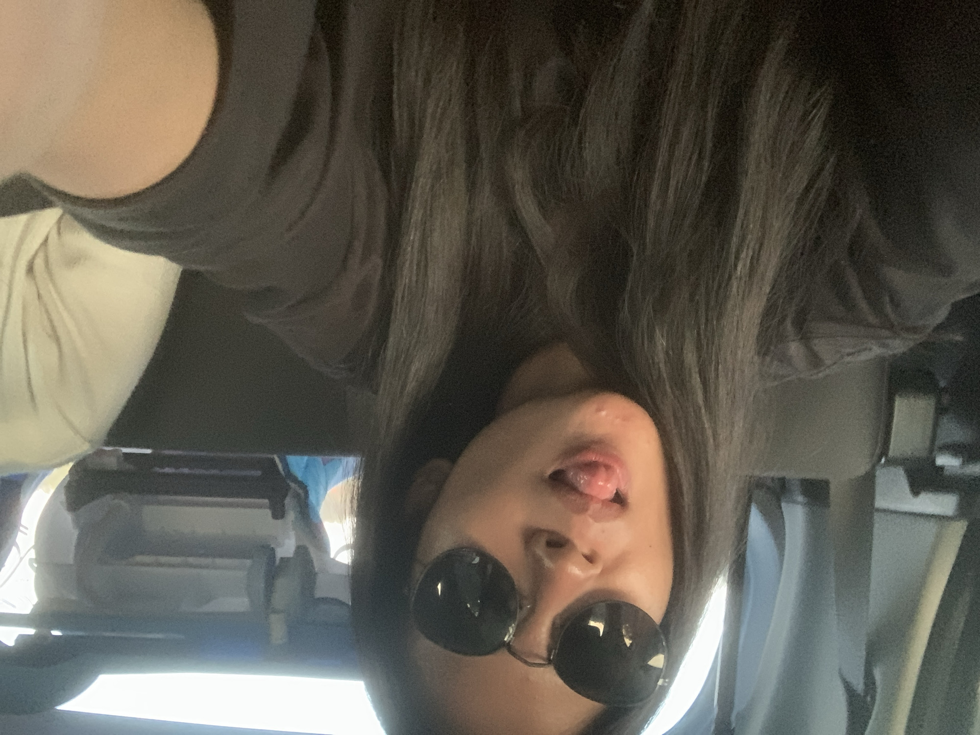 A woman with long, dark hair is taking a selfie from an upside-down perspective. She is wearing black round sunglasses and a black shirt. Her tongue is slightly sticking out. In the background, there is a white headrest and other blurry objects that suggest she might be in a car. The location, Batu Batu, Indonesia is irrelevant to the visual content of the image.