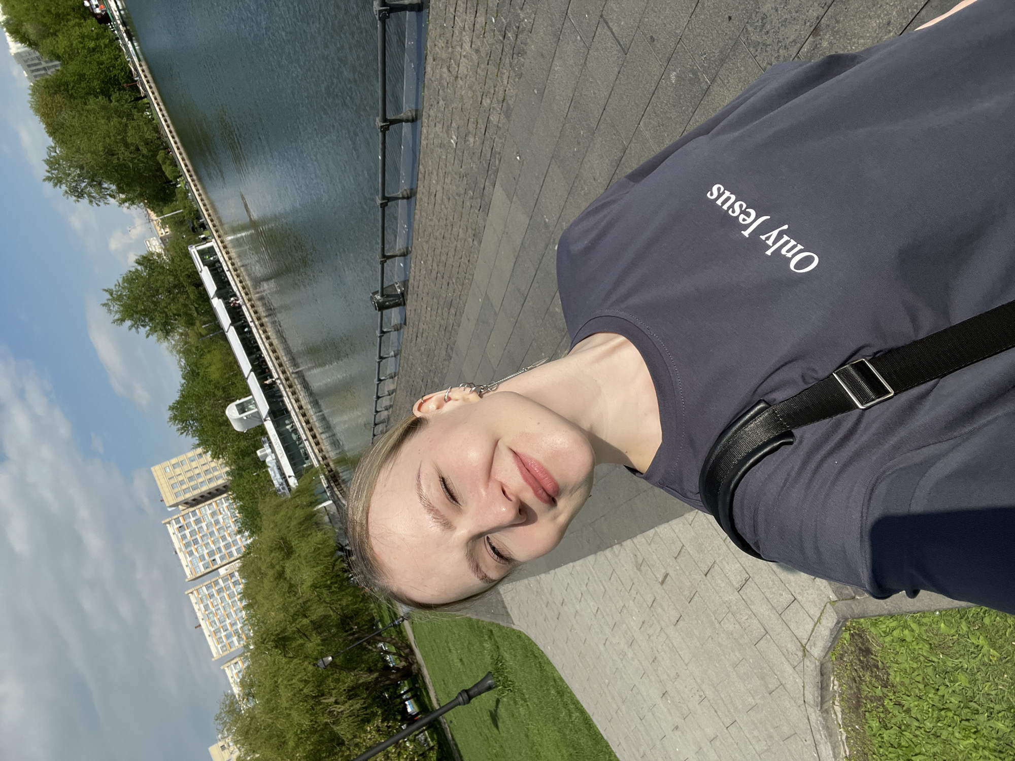 A daytime, tilted, medium-shot captures a fair-skinned woman on a paved walkway along a body of water, potentially the Moscow Canal, in Khimki, Russia. She wears a dark t-shirt with "Only Jesus" printed on the upper left chest and a black shoulder bag strap is visible. Her eyes are closed, and she is facing the camera with a slight smile. In the background are tall residential buildings, green trees, and a cloudy sky. To the left of the walkway is a stretch of green grass and along the waterfront are what appear to be benches and lampposts.