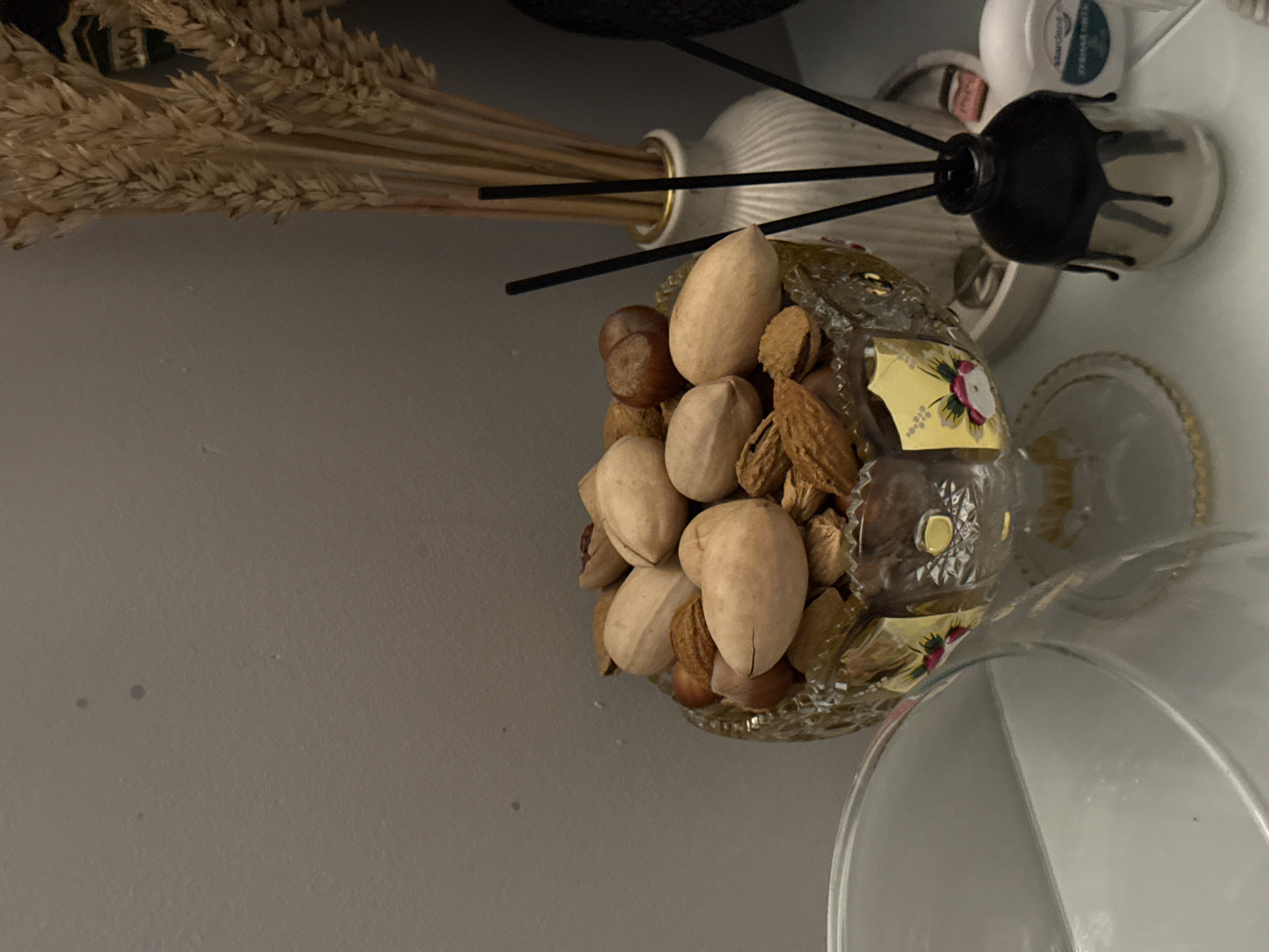 A still life captured from an oblique, top-down perspective shows a decorative composition of various objects placed on a flat, light-colored surface. The objects include a bundle of dried wheat stalks, a small white ceramic vase with thin, black reeds extending upwards, a glass bowl filled with various nuts including pecans, almonds, and hazelnuts, a cylindrical bottle with a dark cap and a narrow neck, a small container of cream, and a clear glass goblet. The goblet and nut-filled glass bowl are intricately designed with floral patterns. The environment is neutral, suggesting a domestic setting. No people are present in the image. Location: Unknown, Unknown.