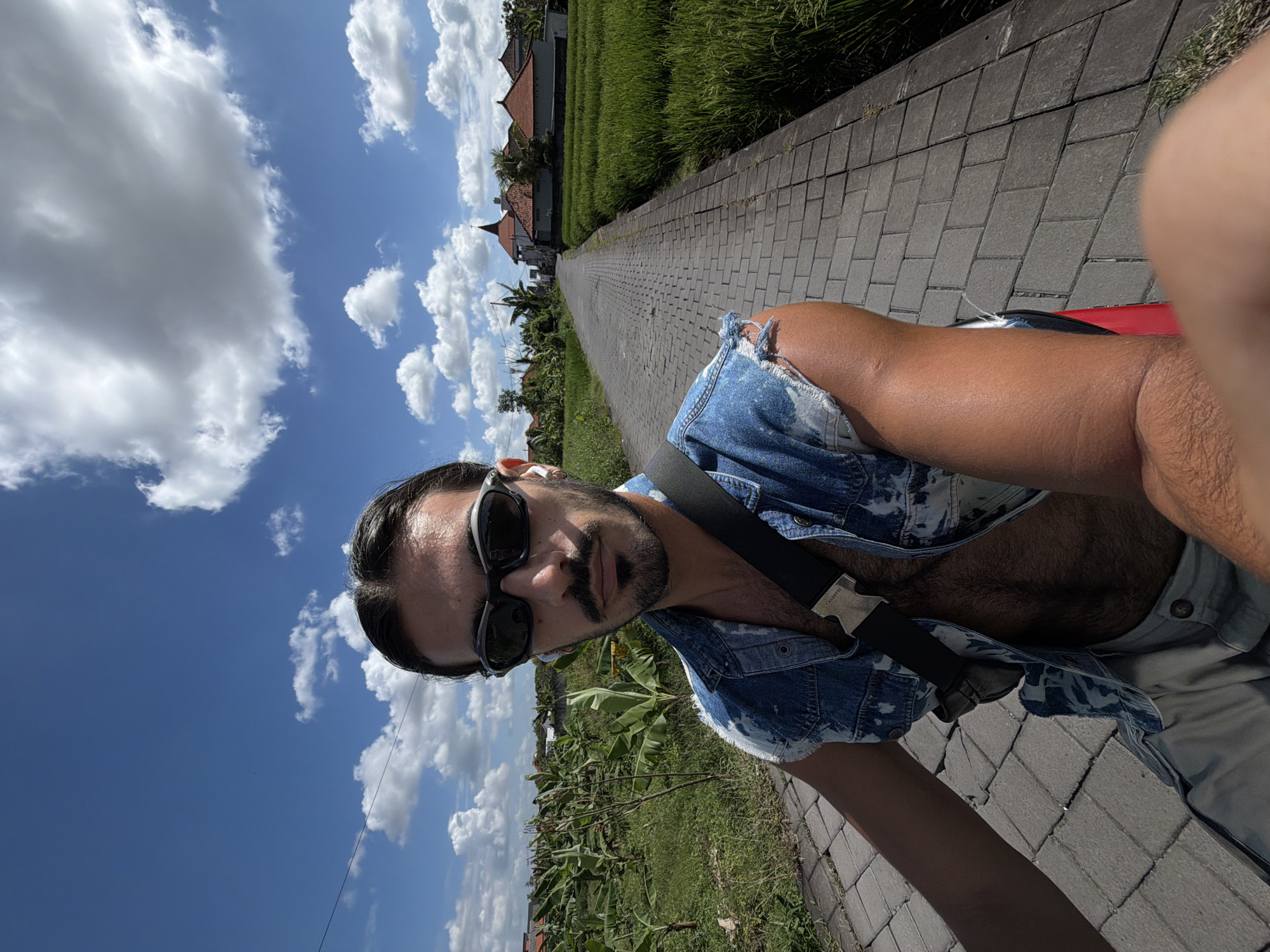 A medium-range selfie-style photograph shows a man with dark hair, a mustache, and sunglasses, wearing a ripped denim vest. A black strap crosses his chest. He is positioned to the right side of the frame, with his head slightly tilted and facing the camera. The man's body is partially visible.
The background features a narrow brick-paved path stretching into the distance, flanked by greenery on both sides. To the left is a blue sky with scattered clouds. On the sides of the path are areas of dense green vegetation. The image appears to have been taken in a natural setting, with possible rice fields visible. A building is visible in the background.
Based on the visual cues in the photograph, it is plausible that this image was taken near rice fields in North Kuta, Indonesia.