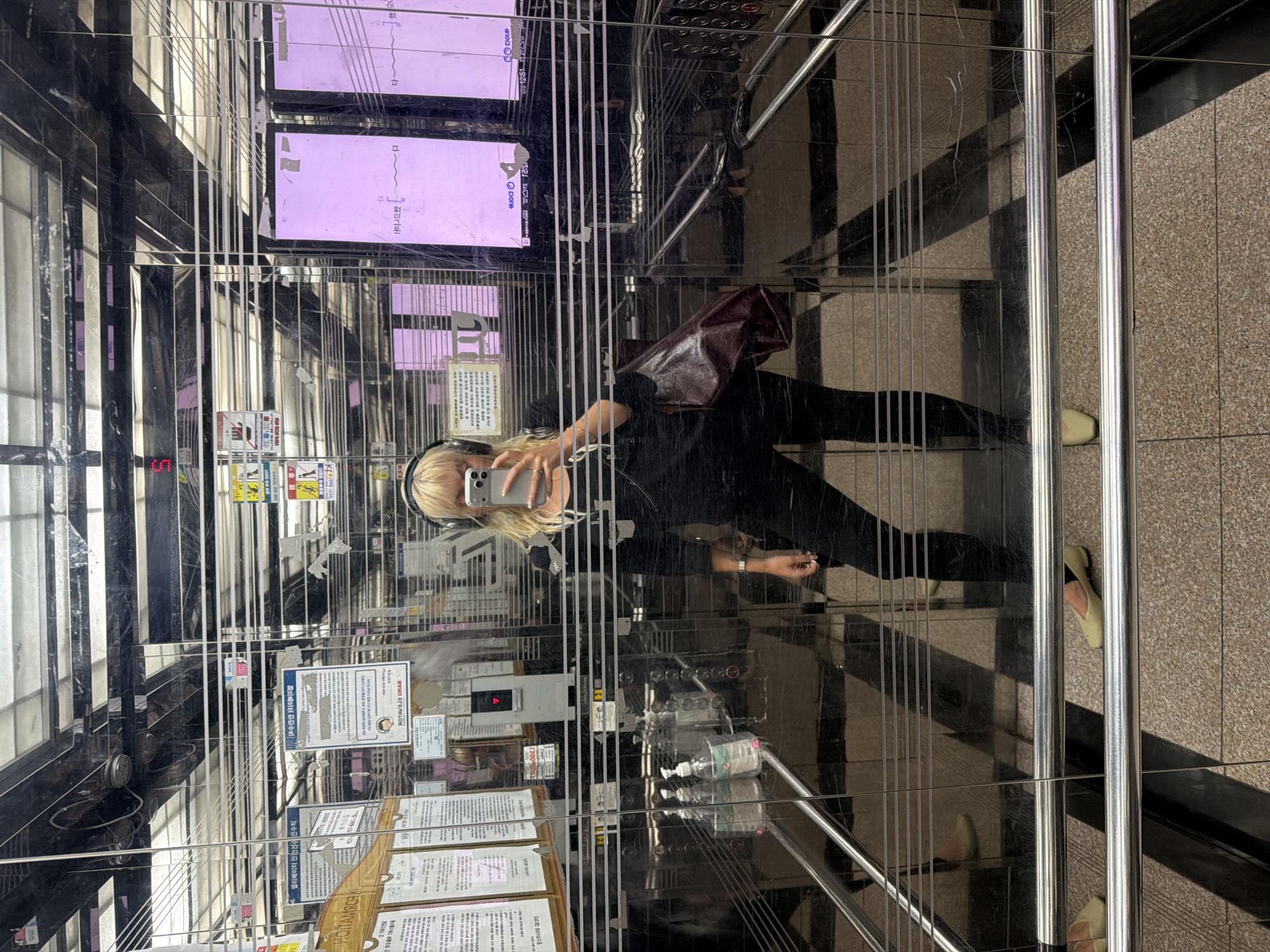 The image captures a person taking a selfie in an elevator. The individual, wearing headphones, black clothing, and light-colored footwear, is holding a smartphone and facing the mirror. Their blond hair is visible. The elevator interior features mirrored surfaces, displaying reflections of the surroundings, including posted notices and signage in Korean. A display screen at the top shows pink information. The elevator has hand sanitizer dispensers. The scene is in Seoul, South Korea, as evidenced by Korean language text in the elevator's signage.