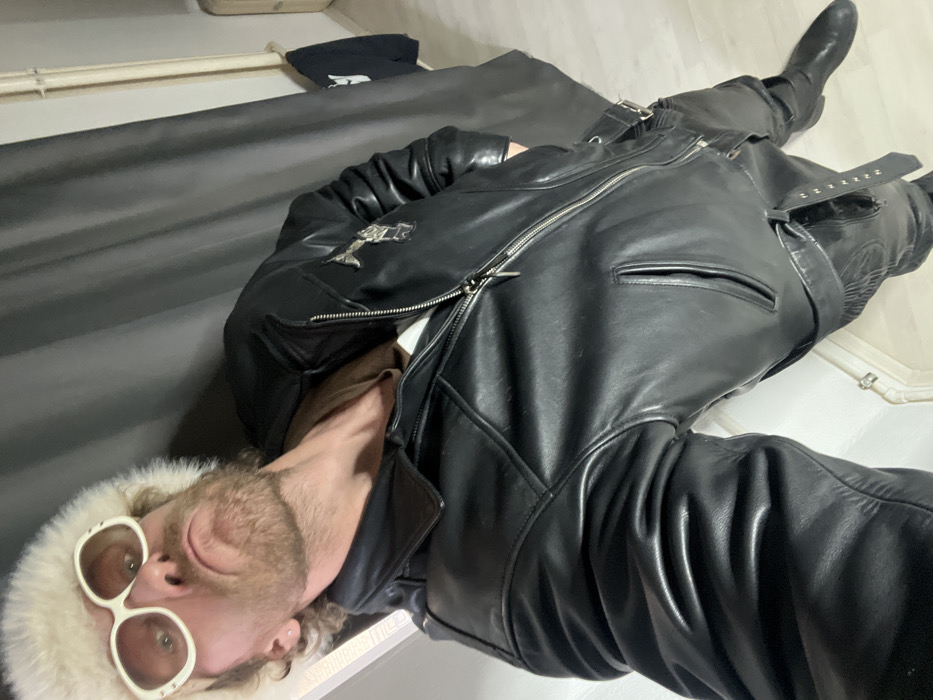 An adult male is depicted in a full-body, low-angle shot within an indoor environment. He is positioned leaning against a light-colored wall or frame on the left, with a dark grey curtain or fabric backdrop on the right. The individual wears a white, fluffy, fur-like ushanka-style hat, large white-framed oval sunglasses with light-tinted lenses, a black leather motorcycle jacket with multiple silver zippers, pockets, and buckles. A silver-colored embellishment, possibly an emblem, is present on the right lapel of the jacket. A plain brown garment is visible beneath the partially unzipped jacket. He also wears black leather pants with straps and buckles, and black leather boots are visible at the bottom right. The man has a beard, mustache, and light-colored curly hair, with a small stud earring in his visible left ear. His facial expression is a deliberate pout with pursed lips. The background includes a light-colored floor, a radiator or heating pipe along the right wall, and a dark garment hanging on a hook further right. The specific location within the city of Saint Petersburg, Russia, cannot be determined from the visual information provided.