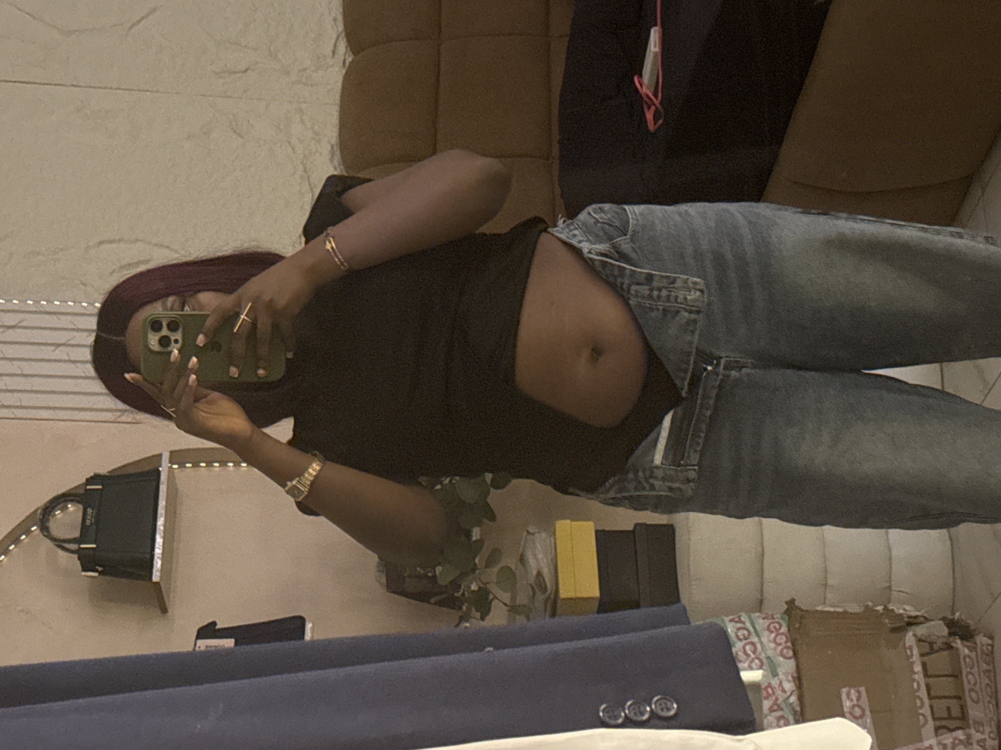 A person is taking a mirror selfie in what appears to be a bedroom in Abuja, Nigeria. The person is wearing a black top and blue jeans, with their stomach exposed. They are holding up a green smartphone to take the picture. In the background, a large brown couch is visible, along with a shelf holding a dark green handbag. There are also stacks of boxes and a blue jacket draped over something to the left of the frame. The lighting in the room is dim, creating a slightly grainy effect on the image.