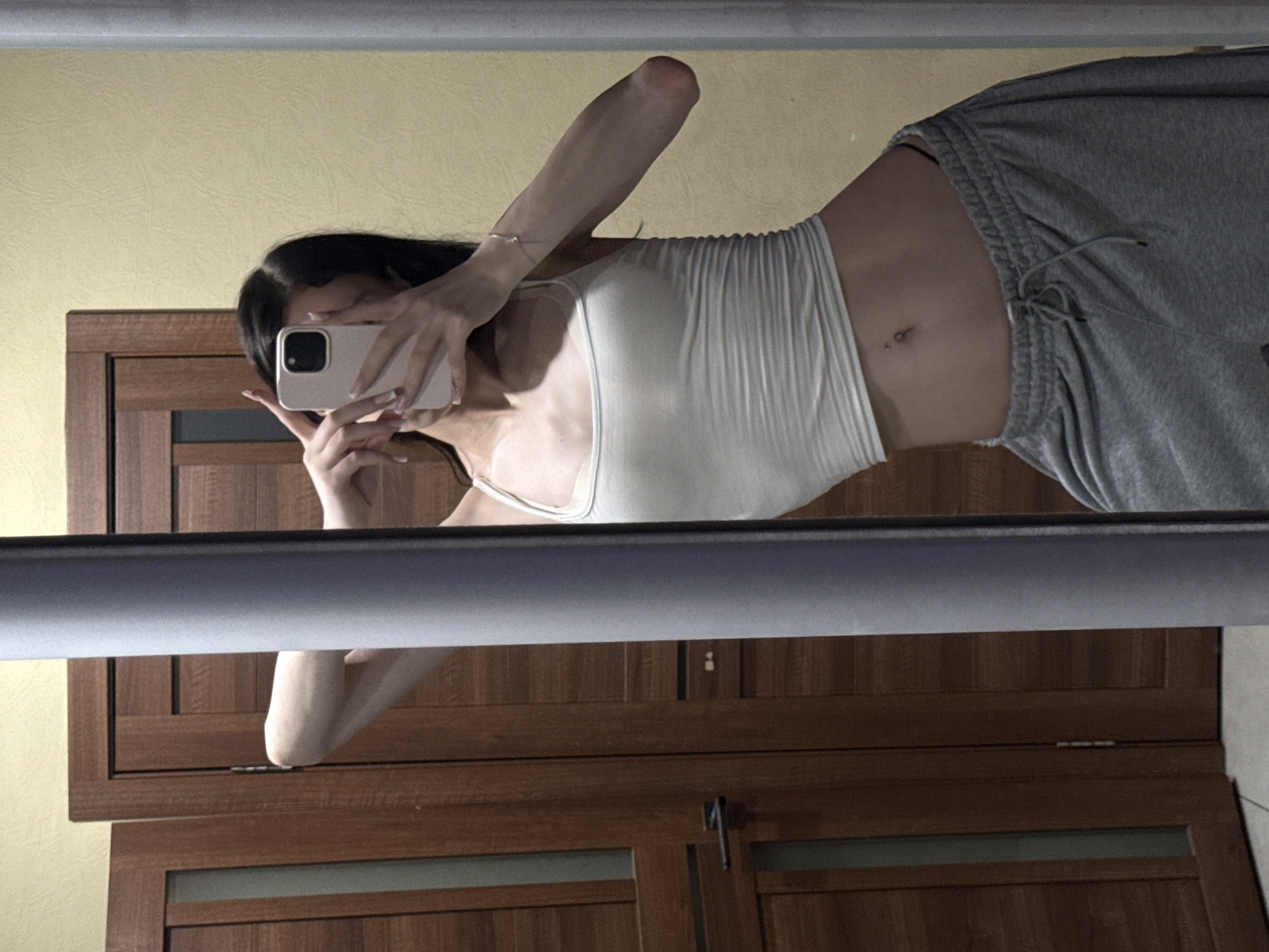 A full-body, mirror selfie shows a young woman with dark hair, wearing a white ribbed tank top and grey sweatpants, posing in an indoor environment. The woman holds a light-colored smartphone in her right hand, obscuring her face, and has her left hand raised near her head, forming a peace sign. A silver bracelet is visible on her left wrist. The background consists of wooden doors and a yellow-textured wall, suggesting a residential or hotel setting. The image appears to have been taken in Khimki, Russia.
