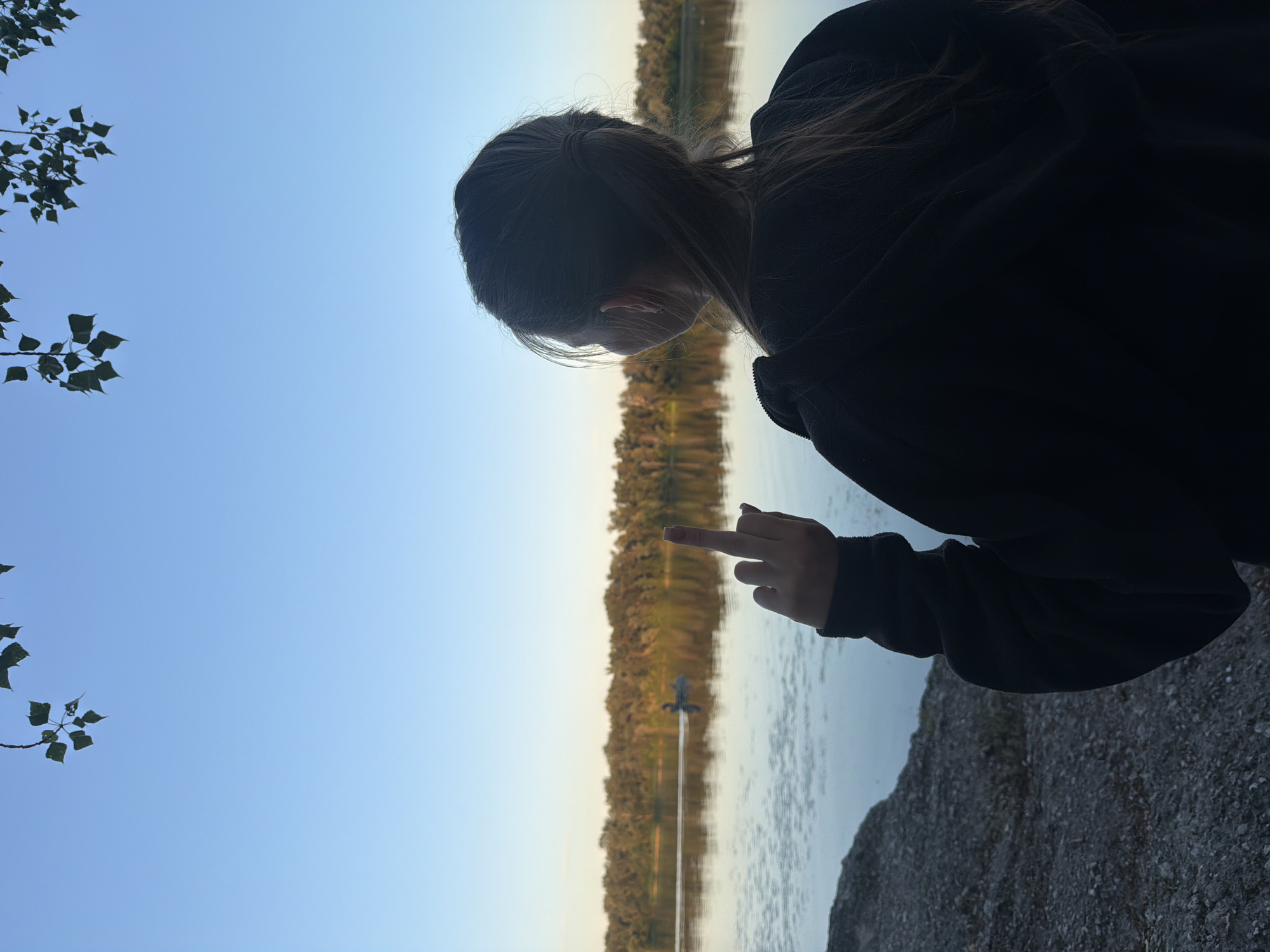 A person wearing a black hoodie stands on a rocky shore, facing a calm river. The person's right hand is raised, with the middle finger extended. The river is bordered by a dense line of trees, which are illuminated by the soft light of a setting sun. The sky is a clear blue, with a few branches and leaves hanging from the top of the frame. In the distance, a small boat with two people is visible on the water. The scene is set in the city of Obukhivka, Ukraine.