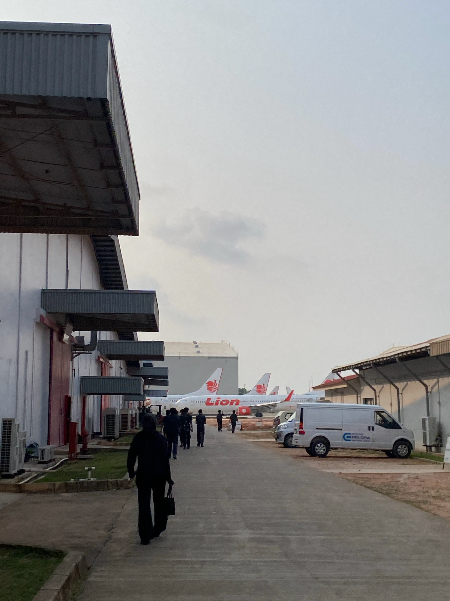 In Batam, Indonesia, a fleet of parked Lion Air airplanes, some with their tails visible, are lined up in an open area. In the foreground, a person in dark clothing walks away from the camera, carrying a dark bag. A white delivery van with the "GELORA" logo is parked to the right. The scene appears to be an airport or aviation maintenance facility, with a large, white building on the left and industrial-style structures on the right. The sky is overcast, and the overall lighting suggests it is either early morning or late afternoon.