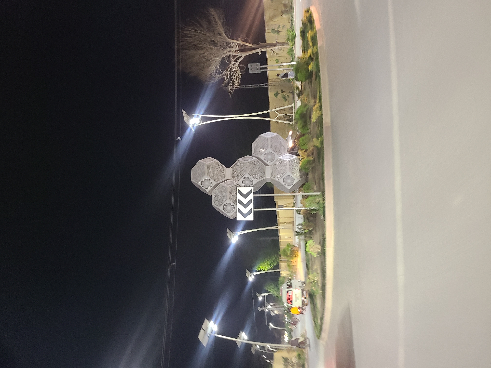 The image depicts a nighttime street scene in Quetta City Tehsil, Pakistan. The sky is dark, indicating late evening or night. Several modern streetlights, each equipped with solar panels, illuminate the area, casting bright white light and visible lens flares.
In the mid-ground, a unique decorative structure stands, composed of multiple interconnected hexagonal units featuring intricate patterns. Below this structure, a white road sign is visible, displaying three black chevron arrows pointing left. Further back, a tall, bare tree with sparse branches is silhouetted against the dark sky.
To the right, a road or lane is present, bordered by a landscaped median or divider planted with green and yellow foliage. A white van and a person on a motorcycle are discernible on the road, suggesting ongoing traffic. Distant buildings or structures are dimly visible beyond the median. The scene highlights modern infrastructure with solar-powered streetlights and decorative elements in an urban setting. No legible text is visible other than the chevron arrows on the sign.