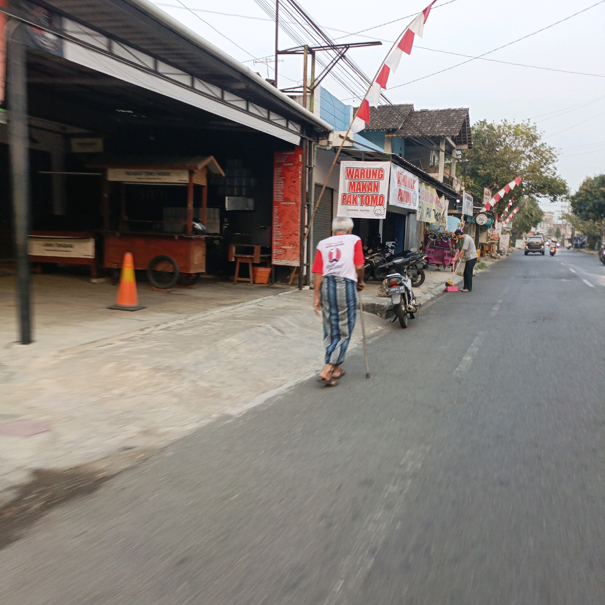 This image depicts a street scene in Salatiga, Indonesia, likely in the afternoon or early evening due to the lighting. In the foreground, an elderly man with gray hair is walking away from the camera, leaning on a walking stick. He is wearing a red and white t-shirt with a logo and a traditional sarong with blue and white stripes. The road surface is asphalt, and to the left, there is a sidewalk or paved area. A bright orange traffic cone with a black base is visible on the sidewalk.
Adjacent to the sidewalk is a row of buildings that appear to be shops or food stalls. One stall has a red and white sign with text that reads "WARUNG MAKAN PAK TOMO" (Pak Tomo's Food Stall), indicating a local eatery. Several motorcycles are parked in front of the stalls. In the background, more buildings with tiled roofs are visible, along with trees and utility poles with numerous wires. Flags, likely Indonesian flags (red and white), are strung across some of the buildings, adding a festive or patriotic touch. Further down the street, a person is sweeping the pavement with a broom. Cars and motorcycles are present on the road, suggesting moderate traffic. The overall impression is of a typical urban street scene in Indonesia.