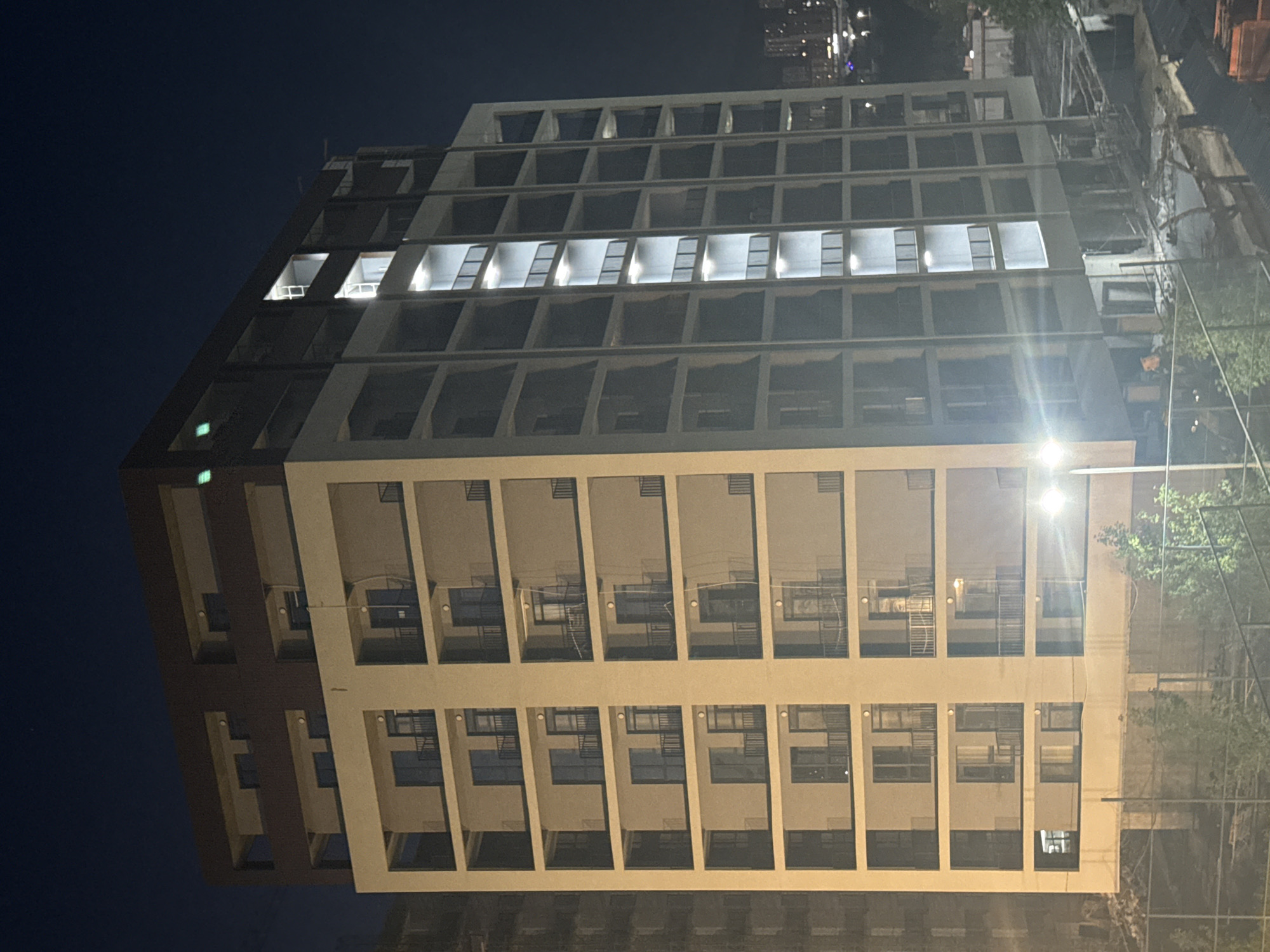 This is a nighttime, low-angle, full-frame photograph of a modern, multi-story apartment building under construction in Tbilisi, Georgia. The building has a light-colored facade with dark balconies and dark window frames. Several floors are visible, each with multiple balcony openings and some windows. Lights are on in some of the upper floors and in the vertical shaft of the building on the right side, illuminating the balconies and windows. The top of the building features darker material, possibly brick or dark cladding, with some visible lights. The ground level of the building appears to be open, with visible structural supports and construction debris. In the foreground, a metal fence runs horizontally, and some trees and bushes are visible. In the background, the lights of the city of Tbilisi are visible, indicating an urban setting. The sky is dark, consistent with nighttime.