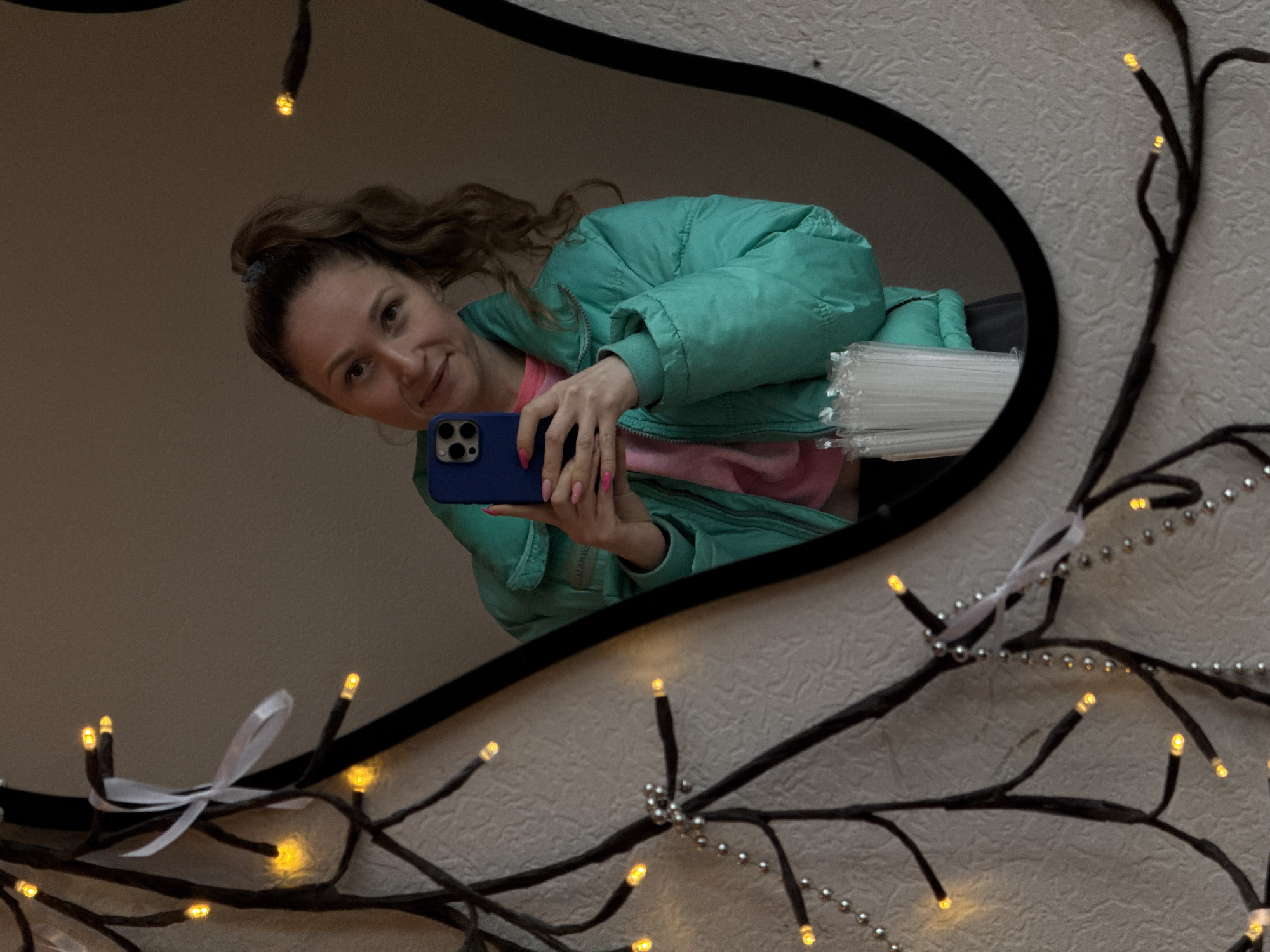 The image shows a woman reflected in a mirror. She is holding a blue smartphone and taking a selfie. She is wearing a light green puffy jacket over a pink shirt. The mirror is decorated with black artificial branches with small white lights. The background wall is light beige with a textured pattern. The scene appears to be indoors. There is no information available to confirm its location in the city of Osa, Russia.