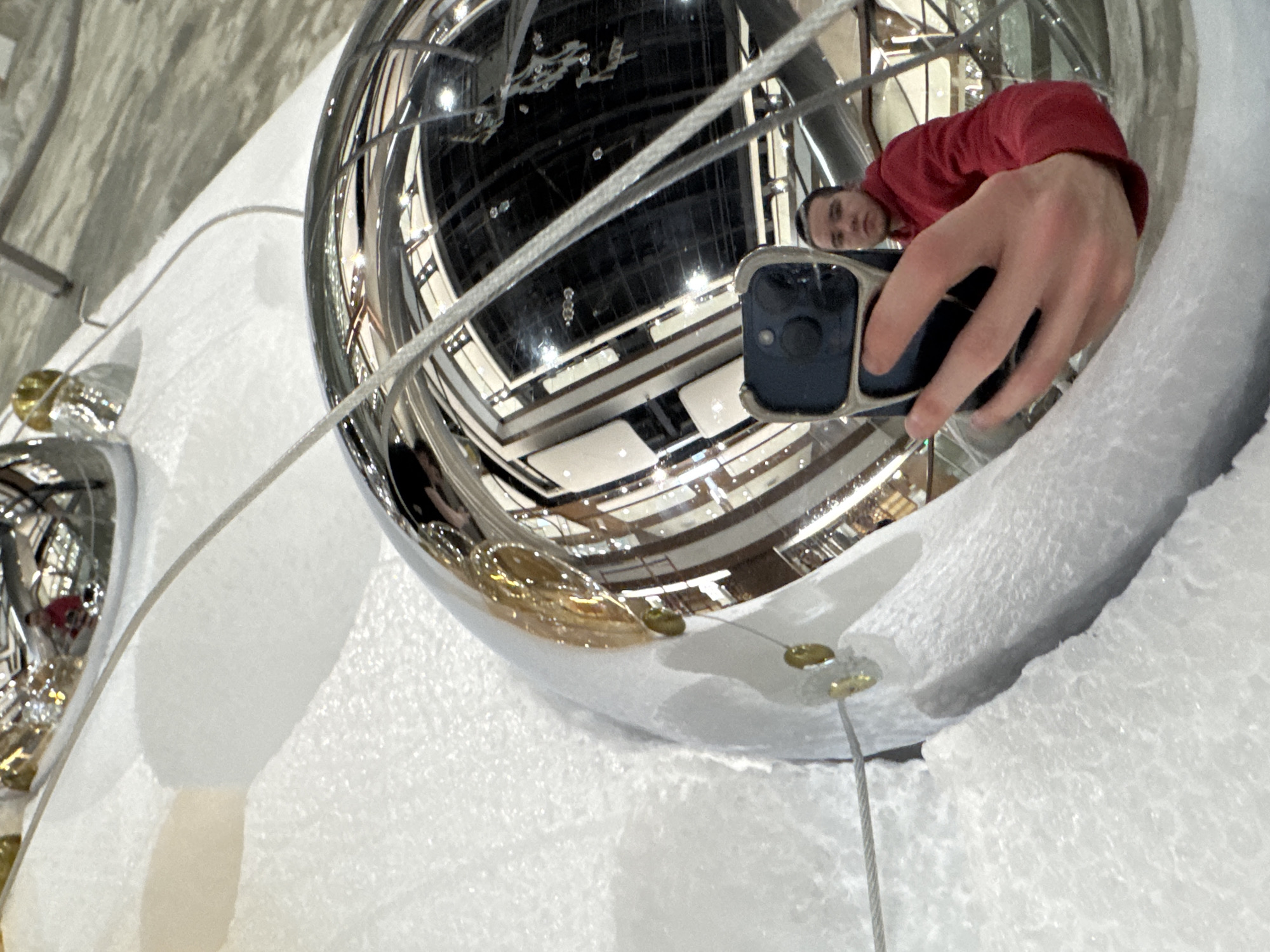 The image captures a person taking a reflection selfie in a large, highly polished spherical ornament. The sphere, appearing to be made of chrome or silver, is partially embedded in or surrounded by white, textured material resembling artificial snow or styrofoam. Thin metallic cables, secured by small gold-colored fasteners, extend from the sphere, suggesting it is part of a larger display or installation.
The reflection in the sphere shows the person, a male wearing a red long-sleeved shirt, holding a dark-colored smartphone to take the picture. His face is visible, looking towards the phone with a neutral expression. The spherical surface distorts the reflection, providing a wide-angle view of the surrounding environment.
This reflected environment is an expansive indoor space, likely a multi-story building such as a shopping mall or atrium, given the visible architectural elements. These include multiple levels with railings, broad structural beams, and a high ceiling with lighting fixtures. The overall scene suggests a festive or decorative display, possibly for a winter holiday, in a public area in Kazan, Russia. No visible text is present in the image.