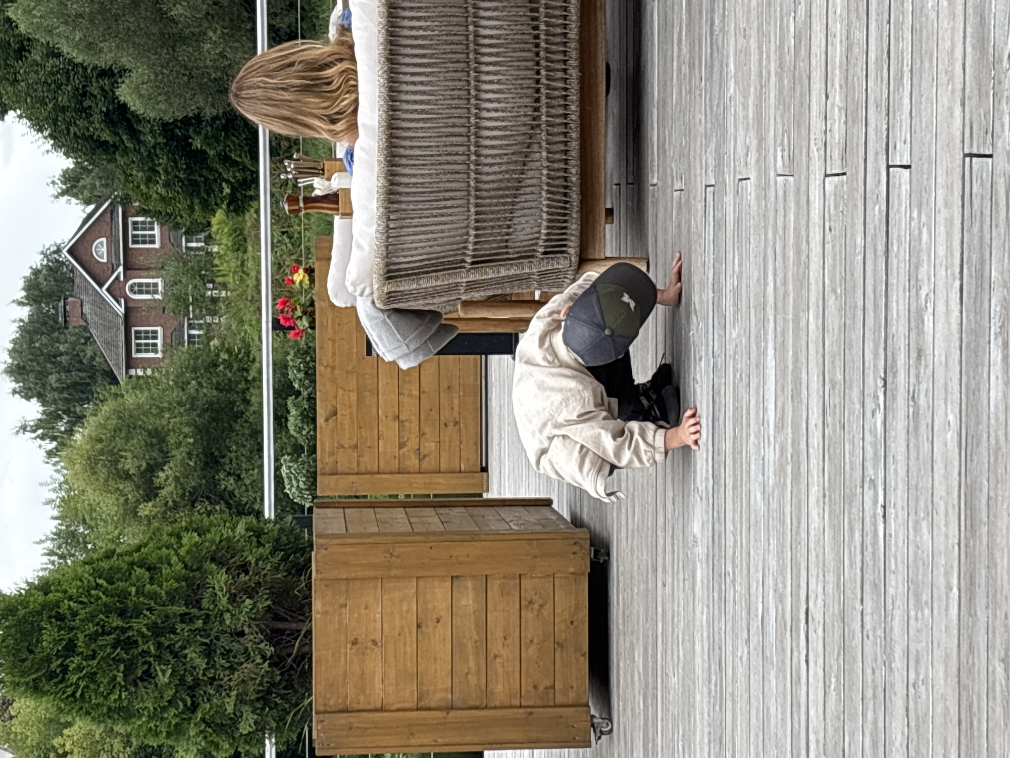 In Zvenigorod, Russia, a young child wearing a light-colored jacket and a baseball cap is crouching on a wooden deck, with their hands on the ground. In the background, a woman with blonde hair is seated on a wicker sofa, facing away from the camera. A red brick house with white-trimmed windows and a gabled roof is visible behind them, surrounded by lush green trees. Two large wooden planters are positioned on the deck, one to the left of the child. The scene appears to be an outdoor dining or lounging area, possibly at a restaurant or residential property.