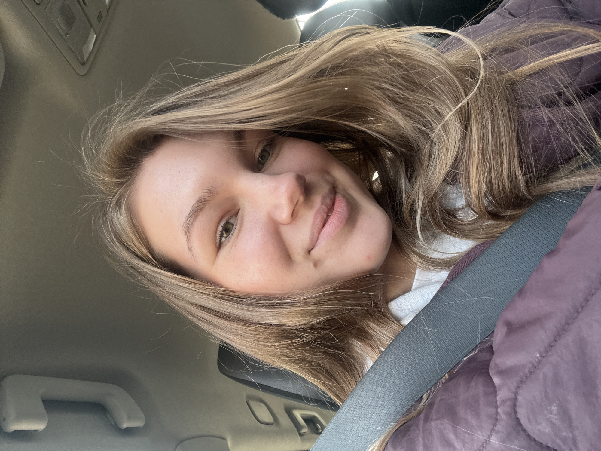 A young woman with light brown hair is taking a selfie in what appears to be the interior of a car. She is wearing a seatbelt. Part of the car's interior, including a grab handle, is visible. She is wearing a white top underneath a maroon jacket. Her facial expression is friendly. There are no elements to confirm a location in the city of Belgrade, Serbia.