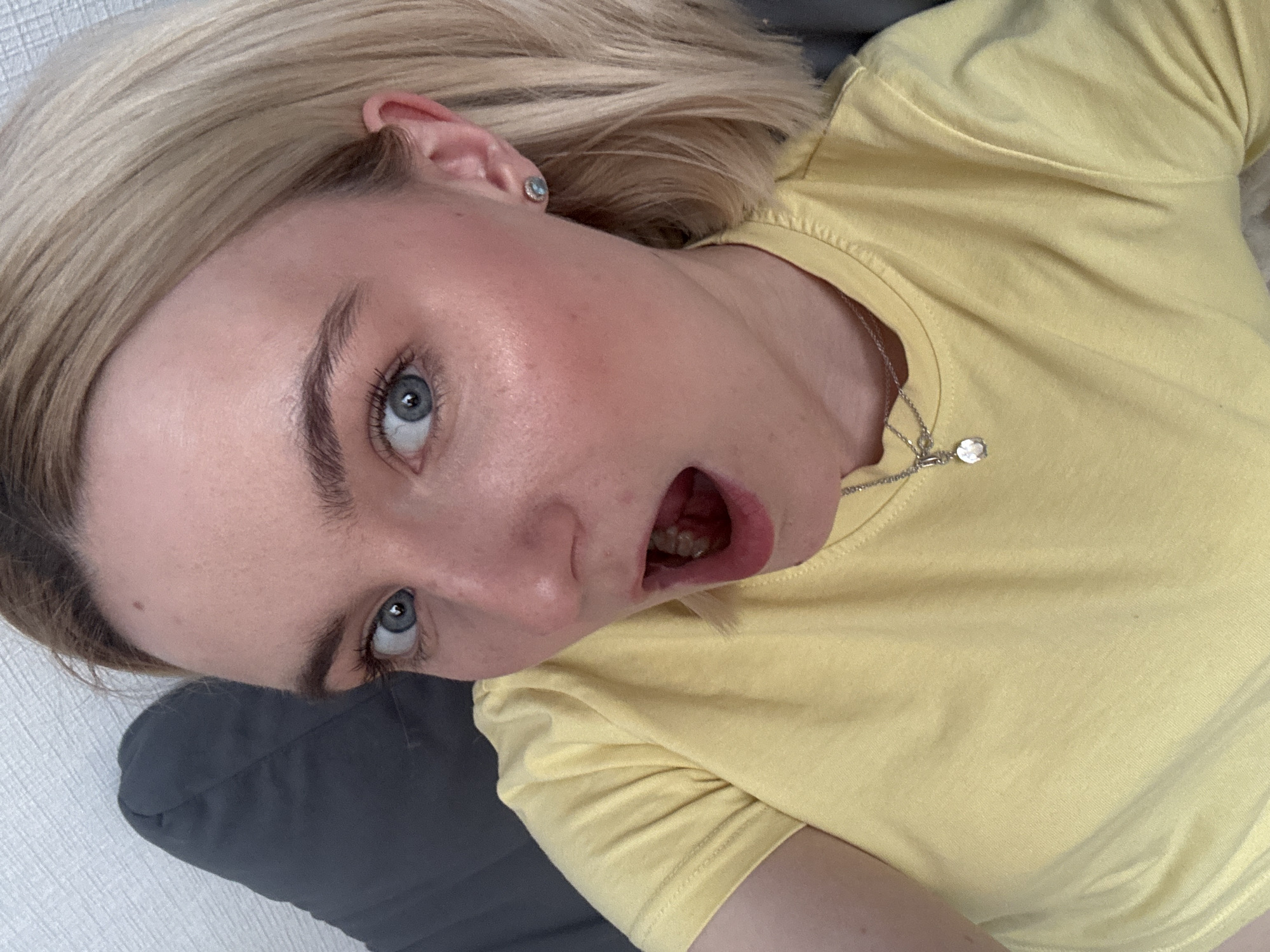 A woman with light blonde hair and blue eyes is lying on what appears to be a grey pillow or cushion. She is wearing a yellow t-shirt and a silver necklace with a round pendant. Her mouth is open in what may be a surprised expression. The location is not evident from the image; there is no information to indicate this photograph was taken in Vsevolozhsk, Russia.