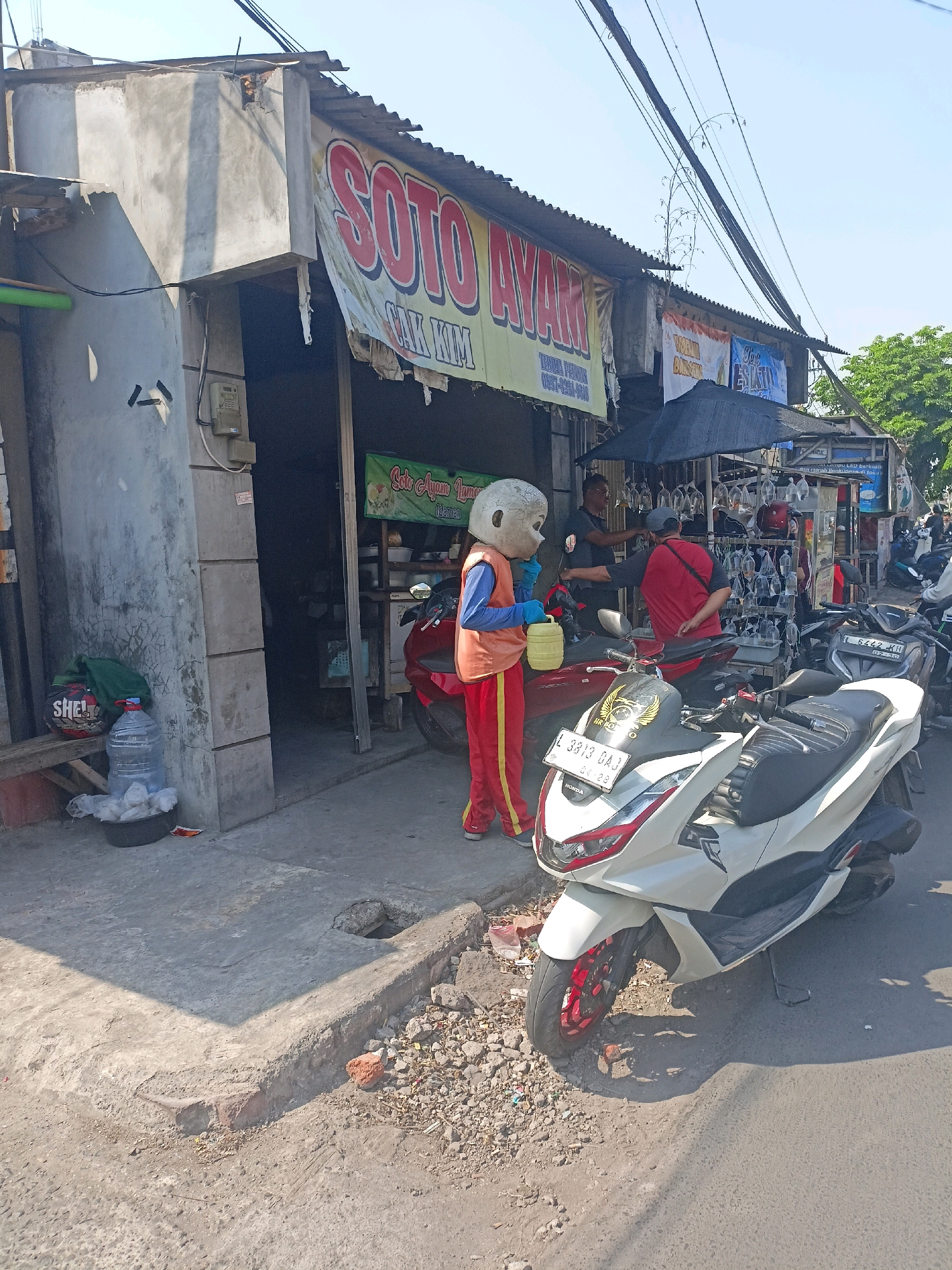 In Surabaya, Indonesia, on a sunny day, a person in a light-colored, round mascot costume is standing on a concrete sidewalk in front of a food stall. The mascot is wearing a blue long-sleeved shirt, an orange vest, red pants with yellow stripes, and yellow gloves. They are holding a yellow water jug with their right hand and have their left hand extended towards it.
The food stall, identified by a banner reading "SOTO AYAM CAK KIM," appears to be a simple structure with a corrugated metal roof. Inside, various items are visible, including what looks like food preparation equipment and possibly seating. Next to the stall, there is a red motorcycle.
In front of the stall and on the sidewalk, several other motorcycles are parked. A white scooter-style motorcycle is prominent in the foreground, with a license plate that reads "L 3813 DAJ". To the left of the mascot, a dark-colored helmet with "SHELL" branding rests on a wooden bench next to a large bottle of water and a black basin filled with white items, possibly plastic bags.
Further back, other people are visible, including two men standing near the stall, one wearing a red shirt and the other in a dark shirt. The environment is characteristic of a bustling street in Indonesia, with overhead power lines and trees visible in the background. The sidewalk is somewhat uneven, with some debris and cracked concrete, and a small hole in the ground. The overall scene depicts a typical street-side commercial area.