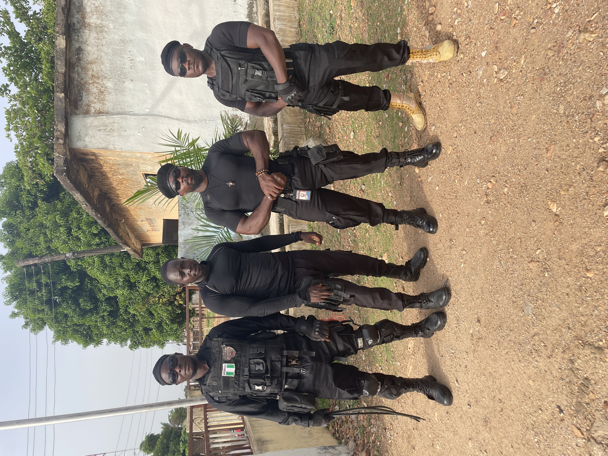 Four men in black tactical gear stand in a line. The first man wears a vest with an identifier reading "NIGERIA." The men wear black boots, except the top man, who wears tan boots. The scene appears to be outdoors on a dirt path with buildings and trees in the background. The location is Akwanga, Nigeria.