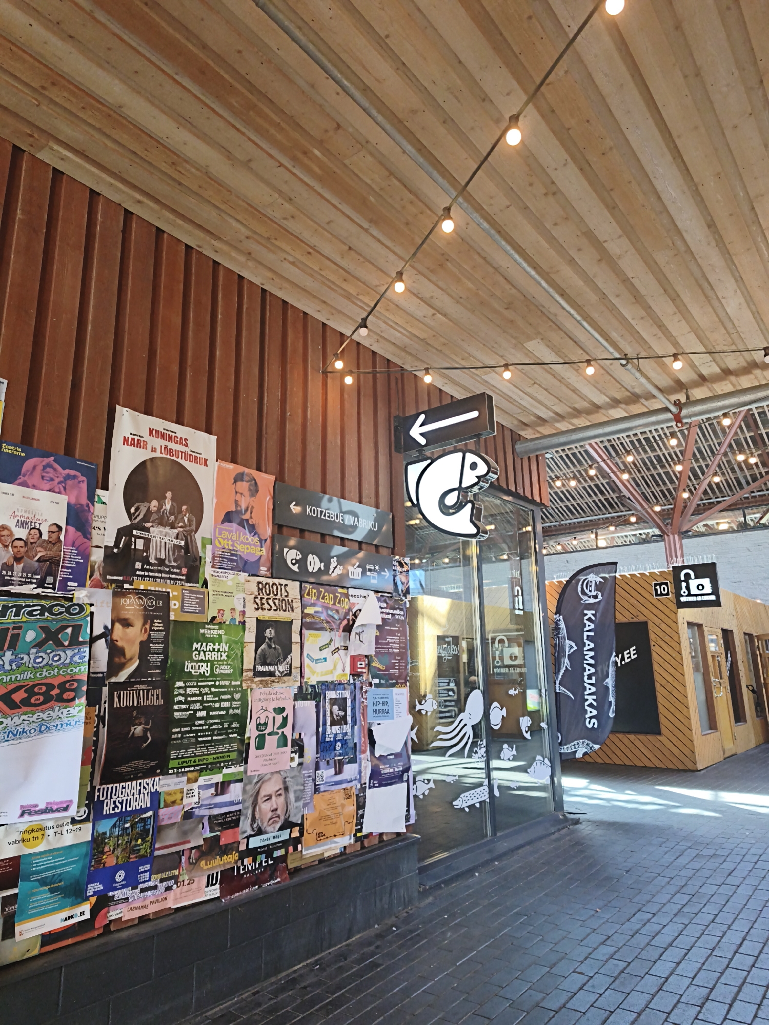 The image shows a street view in the Kalamaja district of Tallinn, Estonia. The scene is dominated by a wall covered in numerous posters and advertisements, suggesting a vibrant artistic or cultural hub. A wooden-paneled building with a glass entrance stands to the right, displaying more signage and a prominent flag with "KALAMAJAKAS" printed on it. String lights hang from the ceiling, illuminating the area. The ground is paved with dark bricks, and a shadow of an overhead structure is cast across it. The posters are varied, featuring text in what appears to be Estonian and images of people, events, and graphics. The overall impression is one of a lively urban environment filled with local cultural information.