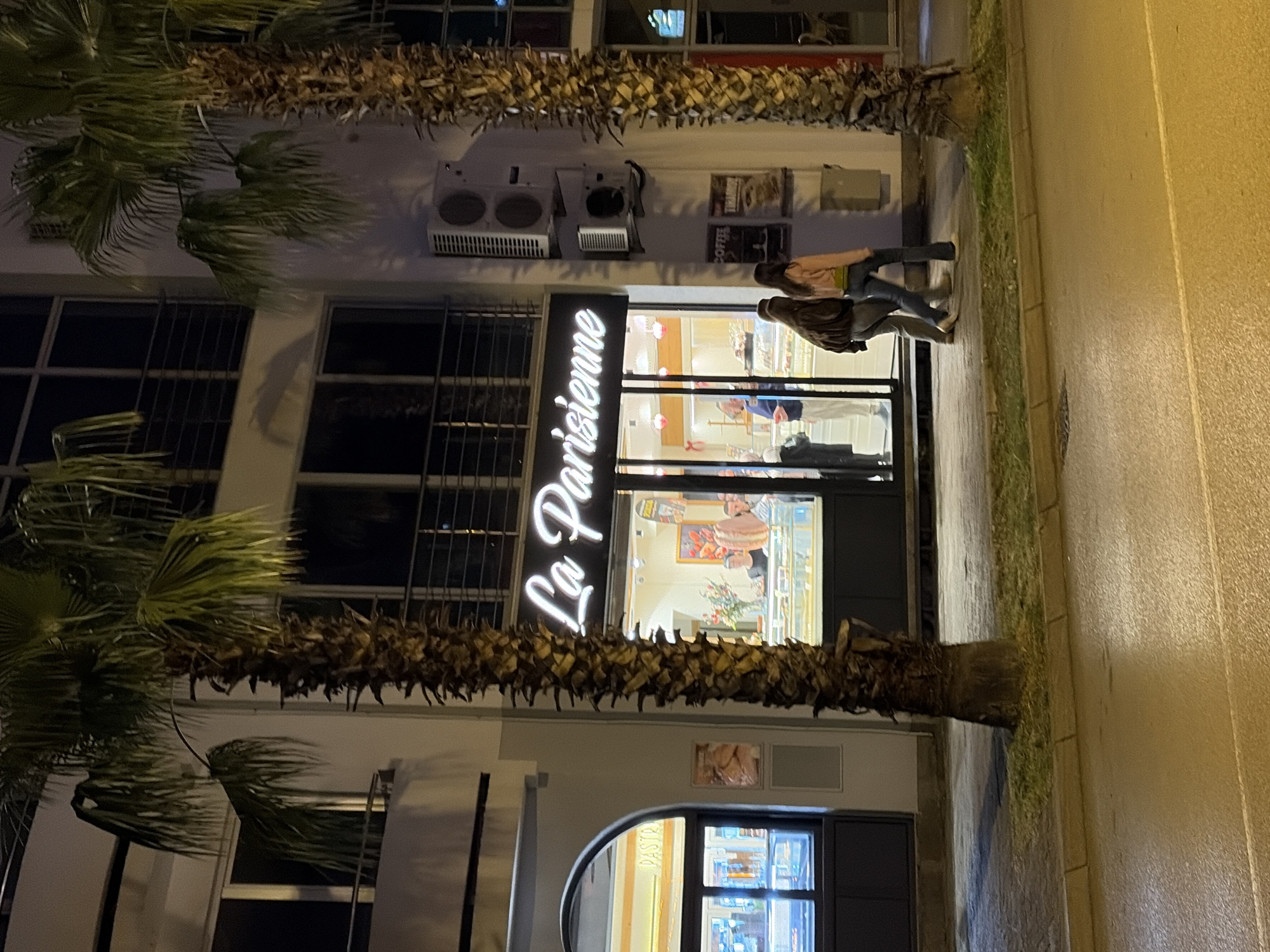 The image shows the storefront of "La Parisienne" in Bar, Montenegro, at night. The storefront features a neon sign with the bakery's name. Two people are walking along the sidewalk in front of the store. Palm trees flank the building. The building also features air conditioning units and framed images on the wall above the lower window.