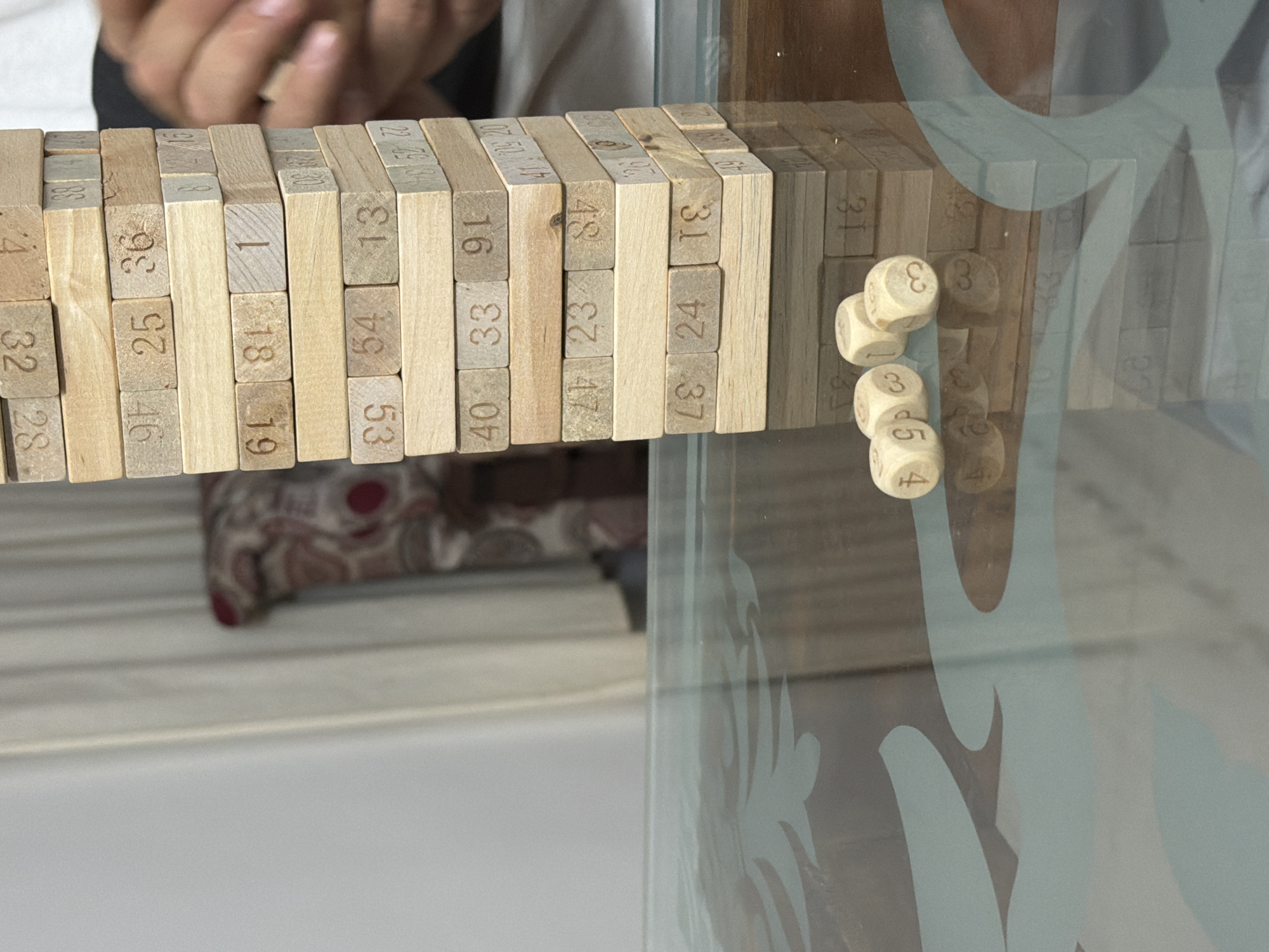 The image depicts a Jenga tower constructed from wooden blocks, each marked with a number. Three dice are scattered in front of the tower on a glass table with a patterned surface. The lighting suggests an indoor setting, possibly a home or a game room. The background shows a curtain and part of a chair, indicating a domestic environment. The overall scene captures a moment during a Jenga game, with the dice suggesting a roll has just occurred or is about to. There is no information in the image that allows for its location to be specified as Thiruvananthapuram, India.