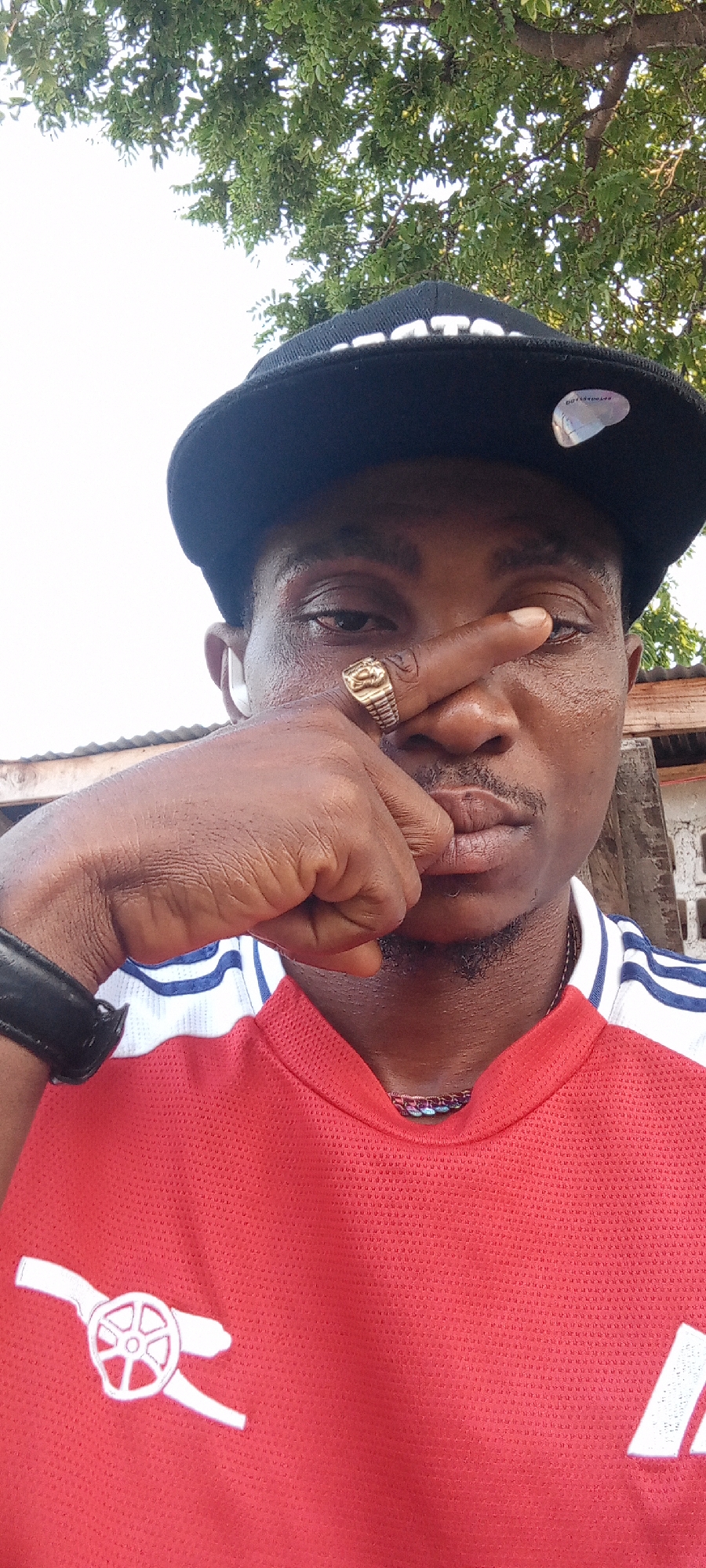 A man is depicted in a close-up shot outdoors, possibly in Zango-ndaji, Nigeria, though the location is not definitively verifiable from the image alone. He is wearing a black baseball cap, a red t-shirt with white stripes on the shoulders, and a large gold ring on his finger. He is holding his hand up to his face, partially obscuring his eye. The background includes foliage and a glimpse of a building.