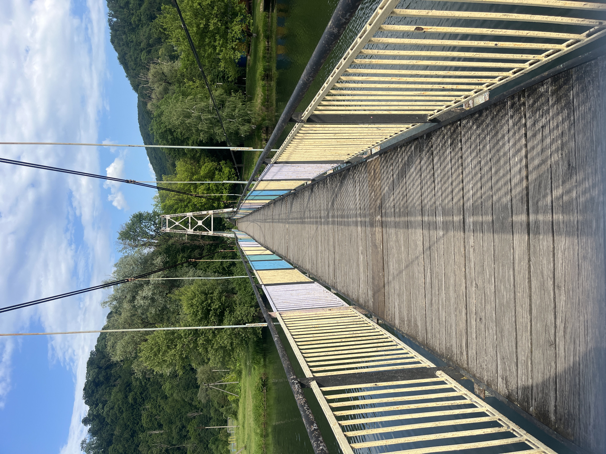 The image captures a wooden suspension bridge stretching across a body of water in Kamianytsia, Ukraine. The perspective is from the bridge deck, looking along its length towards the distant end.
The bridge deck is constructed from weathered, dark wooden planks laid lengthwise. Its side railings feature horizontal metal bars, predominantly painted yellow, with distinct sections of white and light blue appearing further along the bridge, creating a colorful pattern. A black metal handrail runs along the top of these railings. Thin suspension cables are visible overhead, extending from the bridge structure into the bright blue sky. Distinct shadows from the railings are cast diagonally across the wooden deck, indicating bright sunlight. A white metal support tower is visible in the distance, marking a section of the bridge.
Below the bridge, a calm body of water reflects the surrounding environment. Both sides of the water are flanked by steep, densely forested hillsides covered in lush green trees. The sky above is clear blue with scattered white clouds, suggesting a pleasant daytime setting. No people or visible text are present in the image. The scene depicts a serene natural landscape augmented by the man-made structure of the bridge.