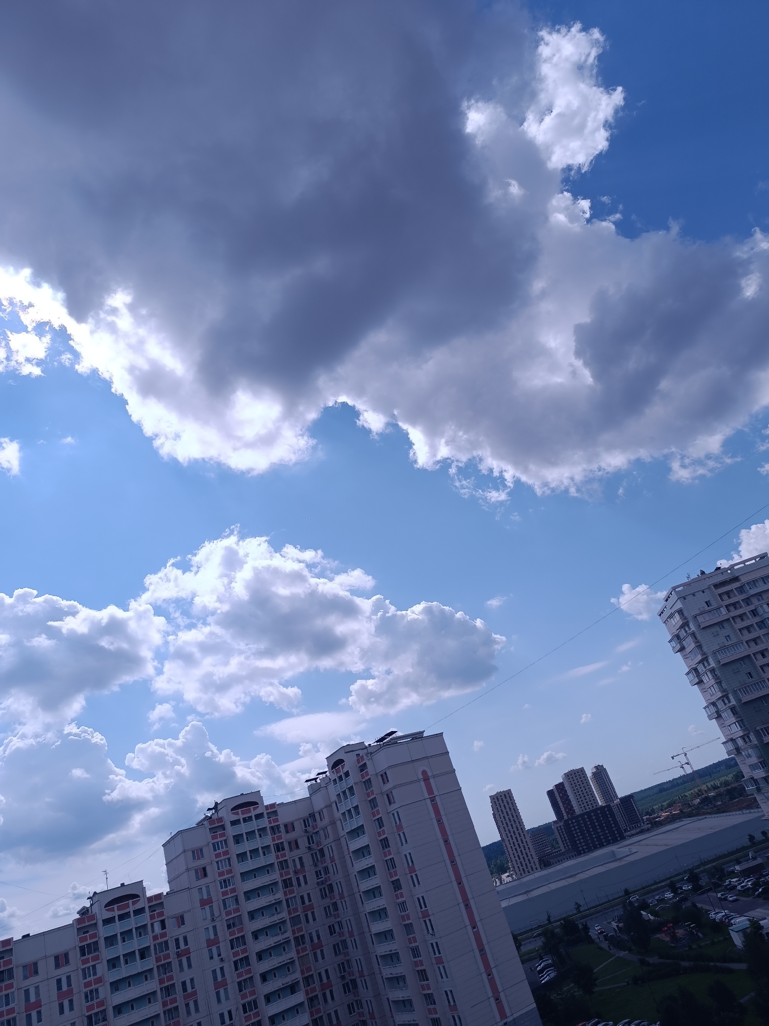 The image captures a city skyline on a cloudy day, with a bright blue sky visible between the clouds. Several high-rise apartment buildings dominate the foreground and mid-ground, characterized by their light-colored facades and numerous windows. Some buildings feature red accents on their balconies and window frames. In the distance, more buildings are clustered together, appearing smaller due to the perspective. The lower part of the image reveals a street with parked cars and some greenery, indicating a developed urban area. The sky is filled with a mix of dark and light clouds, suggesting potential weather changes. No people are discernible in the image.
