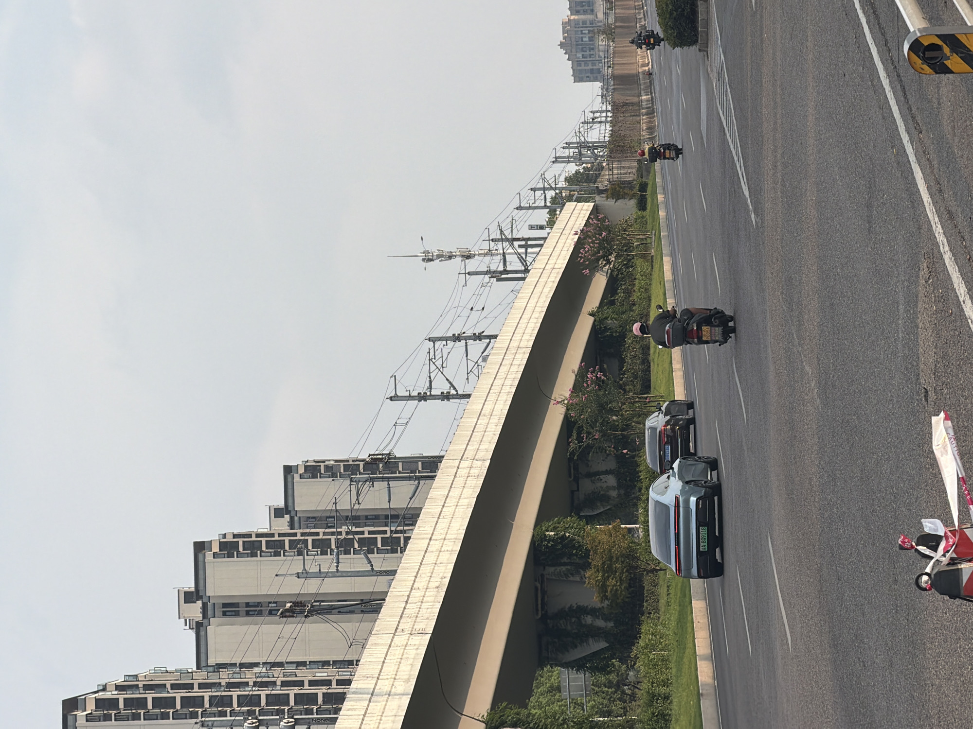 The image captures a street scene in Jiangjin District, China, under a hazy sky. A modern overpass, supported by concrete pillars, dominates the mid-ground, with overhead power lines and a distant telecommunications tower visible. Alongside the overpass, rows of tall, contemporary residential buildings are seen. The road in the foreground is a multi-lane asphalt surface with clear white lane markings. Several vehicles are present: a light blue sedan is in the left lane, followed by a dark sedan in the right lane. Three motorcycles are also on the road; one in the right lane has a rider wearing a pink helmet, another motorcycle further ahead has a rider with a yellow helmet, and a third motorcycle is visible in the distance. Greenery, including bushes and trees, lines the side of the road and the supports of the overpass. In the lower foreground, a red and white object, possibly a traffic cone or safety barrier, is present.