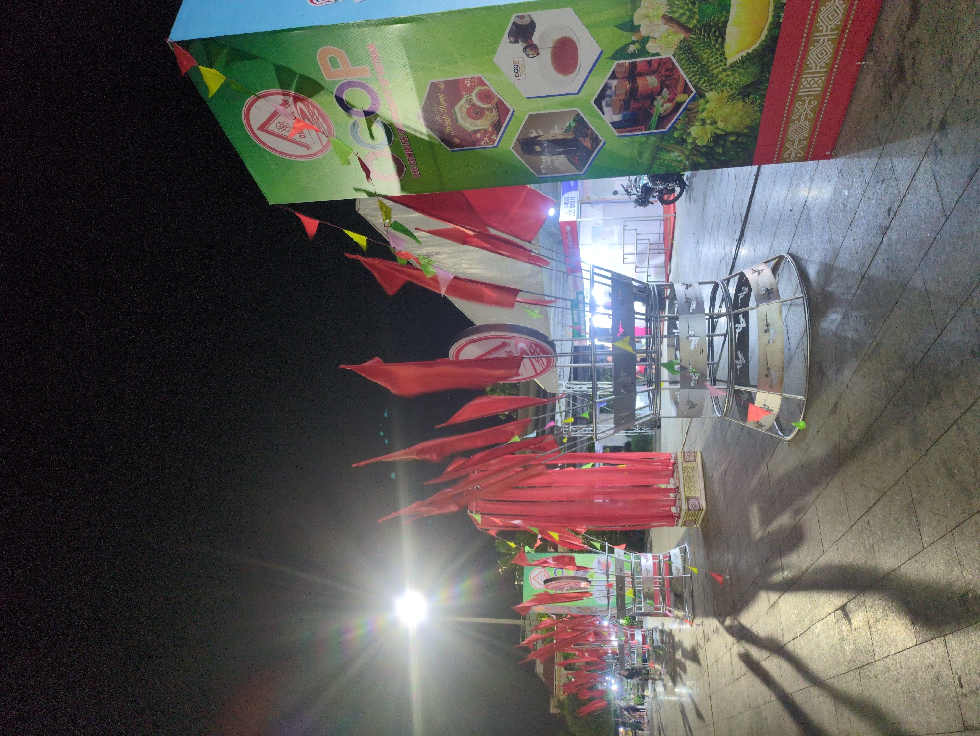 The image shows a street in Tuy Hòa, Vietnam, at night. Rows of metal frames are lined up along the sidewalk, each adorned with red flags. A large banner with the word "COP" printed on it is visible in the background. A streetlight illuminates the scene, casting long shadows. People are walking in the distance. The ground is paved with tiles.
