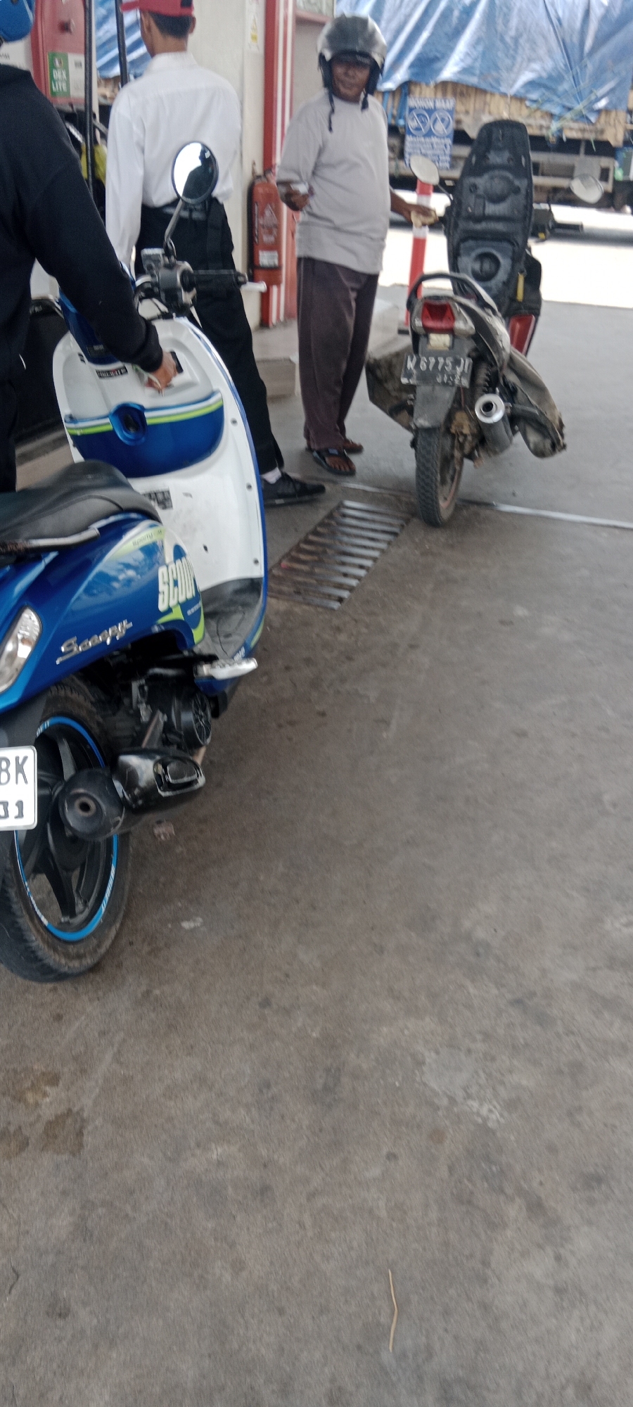 The image depicts a street scene in Tebaloan, Indonesia, likely at a gas station. Two individuals are present. One person, wearing a black jacket and white shirt with a red cap, is partially visible from the left, seemingly interacting with a blue and white scooter. The scooter is positioned prominently in the foreground, its "Scoopy" branding visible on the side. The second individual, dressed in a light-colored t-shirt, dark pants, and a helmet, stands to the right of the blue scooter, holding a mobile phone and looking towards the viewer. This person is standing next to a dark-colored motorcycle with a license plate displaying "N 6775 JD". The environment includes concrete pavement, a grate on the ground, and what appears to be a gas pump structure and possibly a blue tarp in the background, suggesting a commercial area. A red fire extinguisher is also visible on a white pillar to the left. The lighting suggests it is daytime.