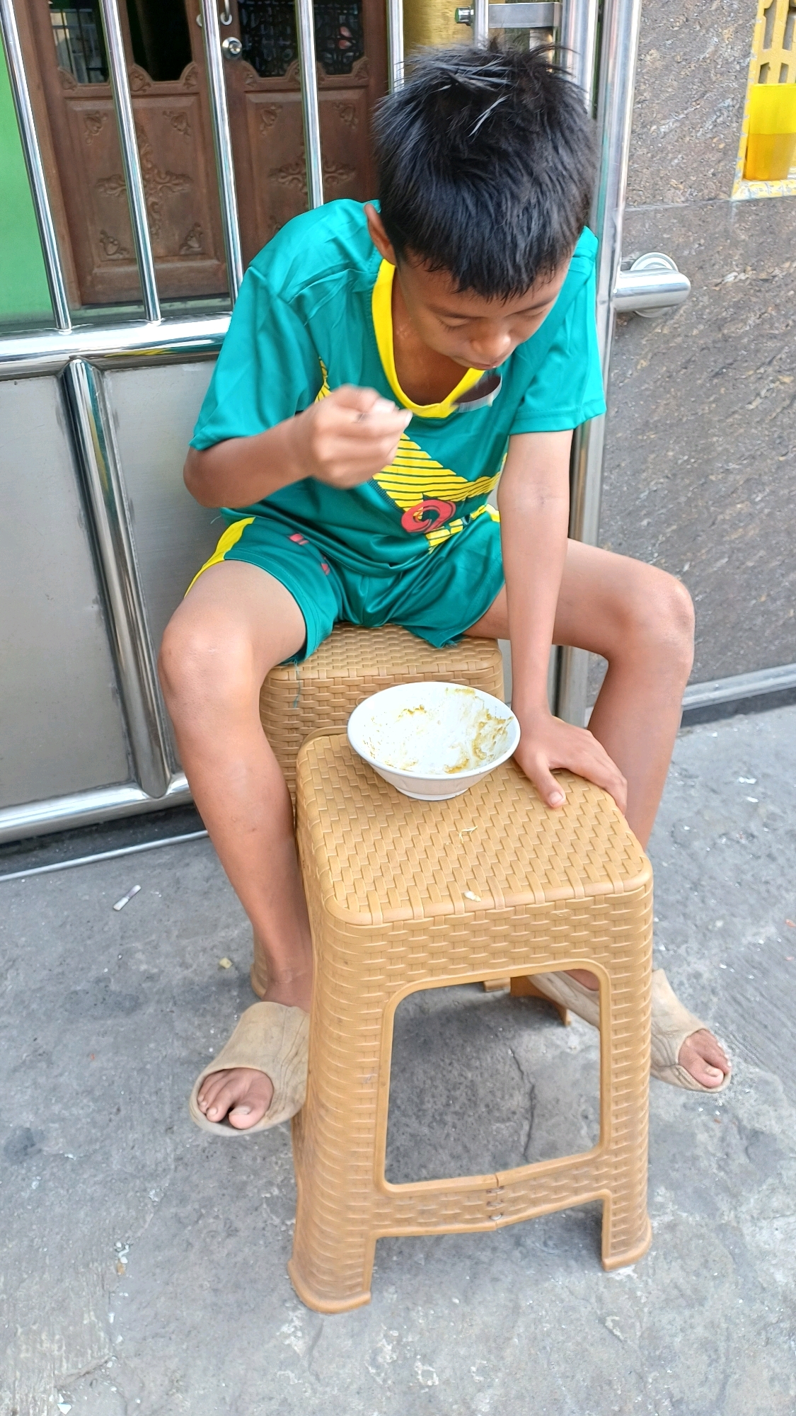 A young boy with dark hair is seated on two stacked plastic stools, eating from a white bowl with a spoon. He is wearing a teal and yellow t-shirt and green shorts, with beige slippers on his feet. The stools are placed on a concrete surface, likely an outdoor area. In the background, there is a metal gate and a wooden door, suggesting a residential or commercial setting. The image appears to have been taken in Citeureup, Indonesia, a suburban area known for its industrial presence.