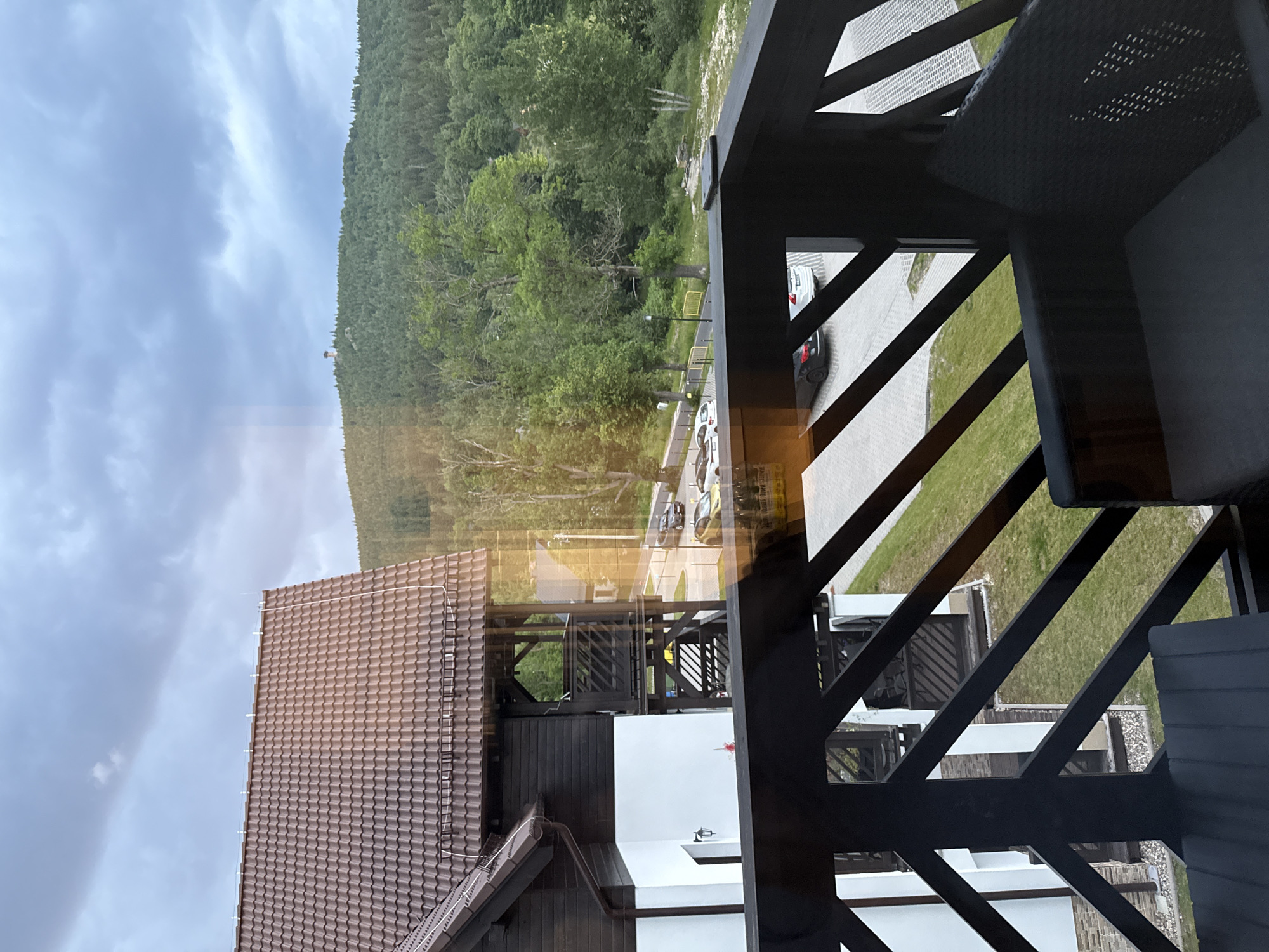 The image shows a view from a balcony overlooking a wooded hillside with a small tower on its summit. In the foreground, a dark wooden railing frames the scene. To the left, part of a building with a brown tiled roof, dark wood siding, and white walls is visible. Below, a parking area with several cars is situated near the building. The overall impression is a serene, natural setting with a modern accommodation. This scene is consistent with the landscape surrounding the region of Szklarska Poręba, Poland, known for its mountainous terrain and forested areas.