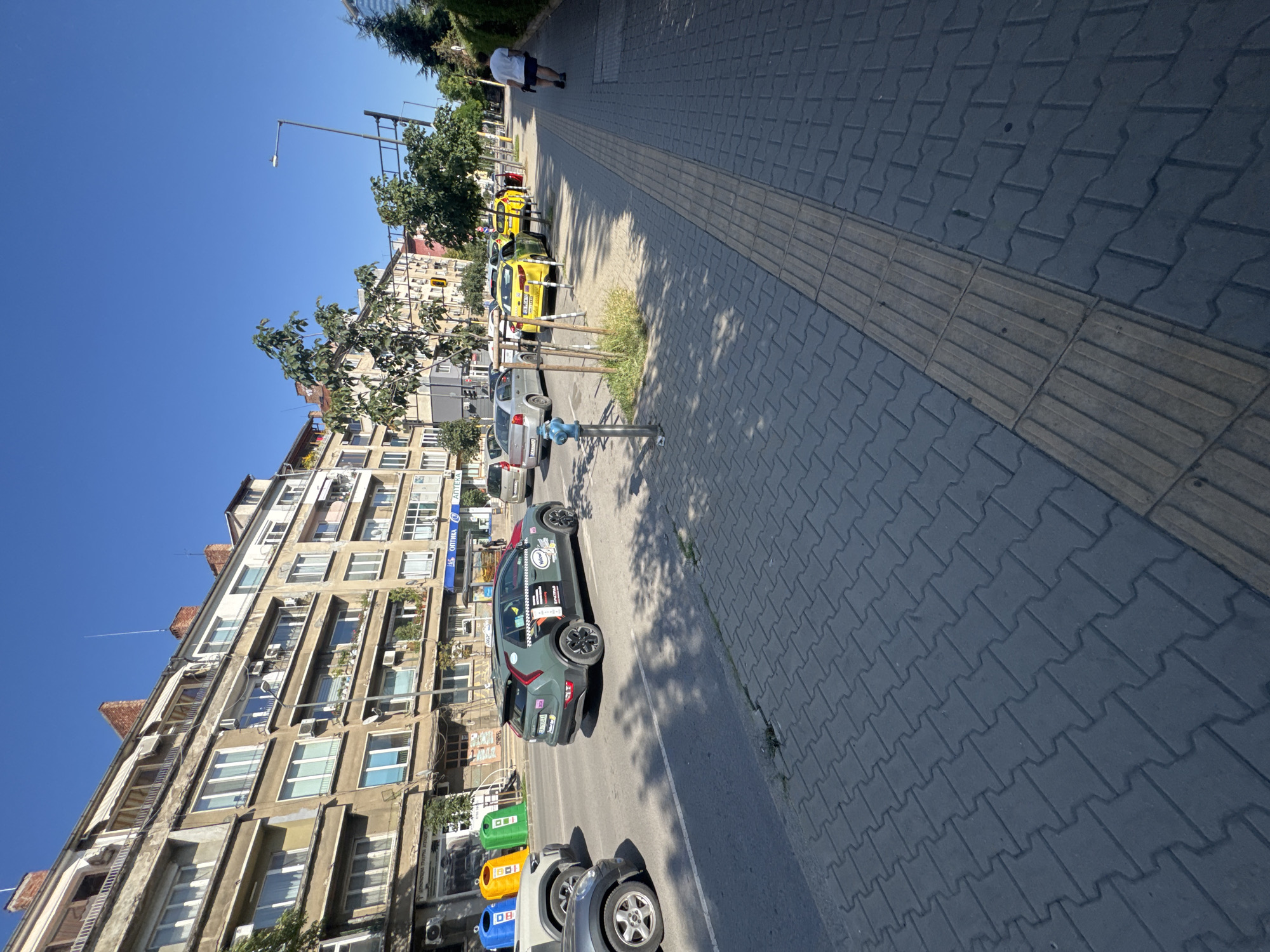 The image shows a street scene in Sofia, Bulgaria, on a bright, sunny day. The perspective is tilted, giving a slightly unusual view of the urban environment.
In the foreground, a textured, grey paved sidewalk runs alongside a street. This sidewalk features a tactile paving strip, designed for visually impaired pedestrians. A blue fire hydrant is visible on the sidewalk.
The street itself is lined with parked cars and a few trash bins. Notably, there is a grey electric car with various stickers, possibly a ride-sharing vehicle, parked near the sidewalk. Further down the street, several yellow taxis are lined up. In the background, a multi-story apartment building with numerous windows and air conditioning units dominates the left side of the frame. Trees and other buildings are also visible, along with traffic lights. A person in a white shirt and dark shorts is walking on the sidewalk in the distance.
The scene appears to be set during daytime, with clear blue skies and bright sunlight casting shadows, indicating fair weather. The presence of taxis and apartment buildings suggests a residential or mixed-use urban area. Some text is visible on the building in the background, including what appears to be "Оптика" (Optika, meaning Optics) and "Аптека" (Apteka, meaning Pharmacy).
