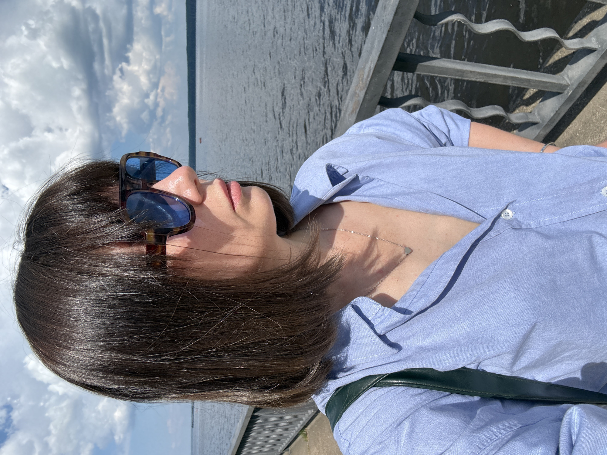 A medium shot shows a woman with dark brown hair and bangs wearing sunglasses and a light blue collared shirt. She is standing next to a metal railing with a body of water and a cloudy sky in the background. The scene appears to be a waterfront promenade in the city of Chaikovsky, Russia.