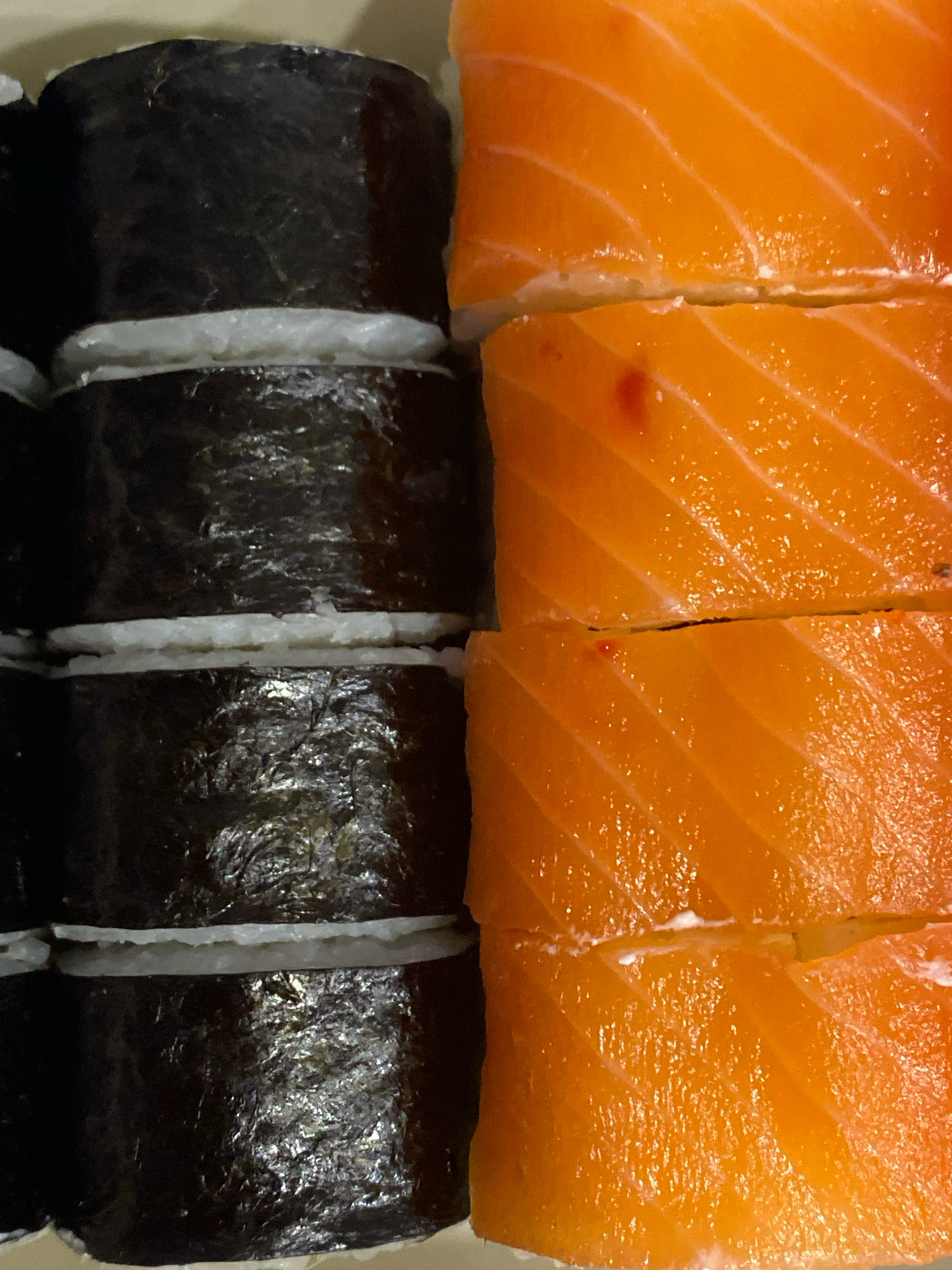The image is a close-up, overhead shot of a platter of sushi rolls. On the left side of the image are several sushi rolls made with dark seaweed and white rice. On the right side are slices of salmon, layered to create a vibrant orange hue with white marbling. The lighting is bright and even, suggesting an indoor setting. There are no people or specific city landmarks visible. The location is not Izhevsk, Russia.