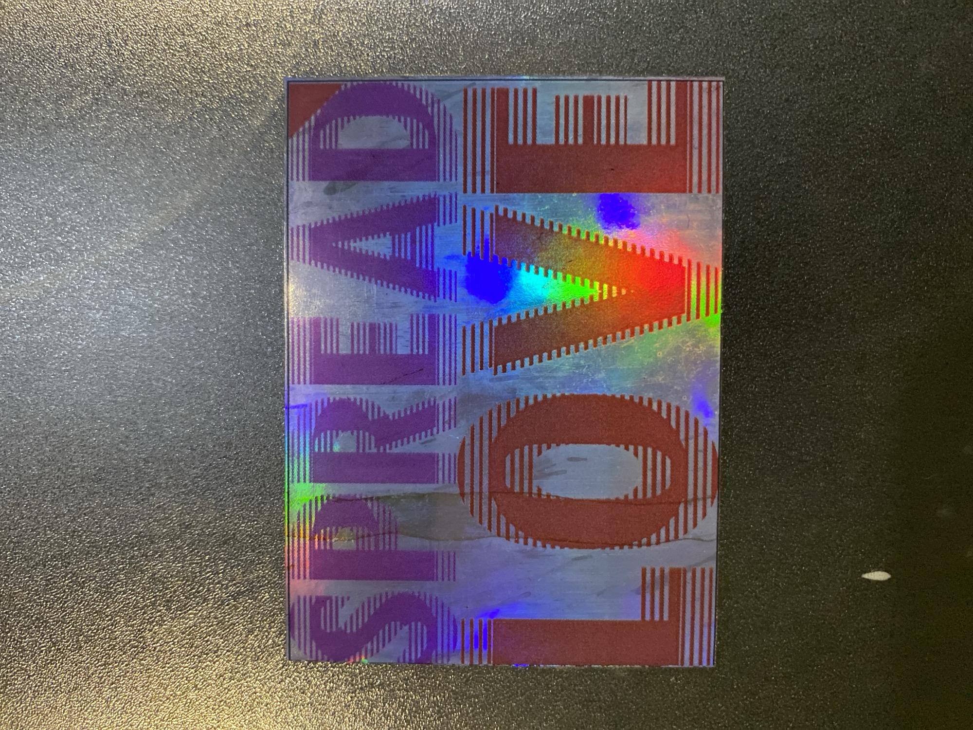 A holographic sticker displaying the words "SPREAD LOVE" is placed on a textured dark surface. The sticker is rectangular and predominantly features purple lettering for "SPREAD" and red lettering for "LOVE." The holographic effect creates a rainbow of colors across the sticker, particularly visible within the letters and the background. The surface the sticker is on appears to be a table or countertop with a coarse, matte finish. There are no people or other objects visible in the immediate vicinity of the sticker. The lighting suggests an indoor environment, possibly a workspace or a home. The location in Kathmandu Metropolitan City, Nepal, cannot be determined from the visual information provided.