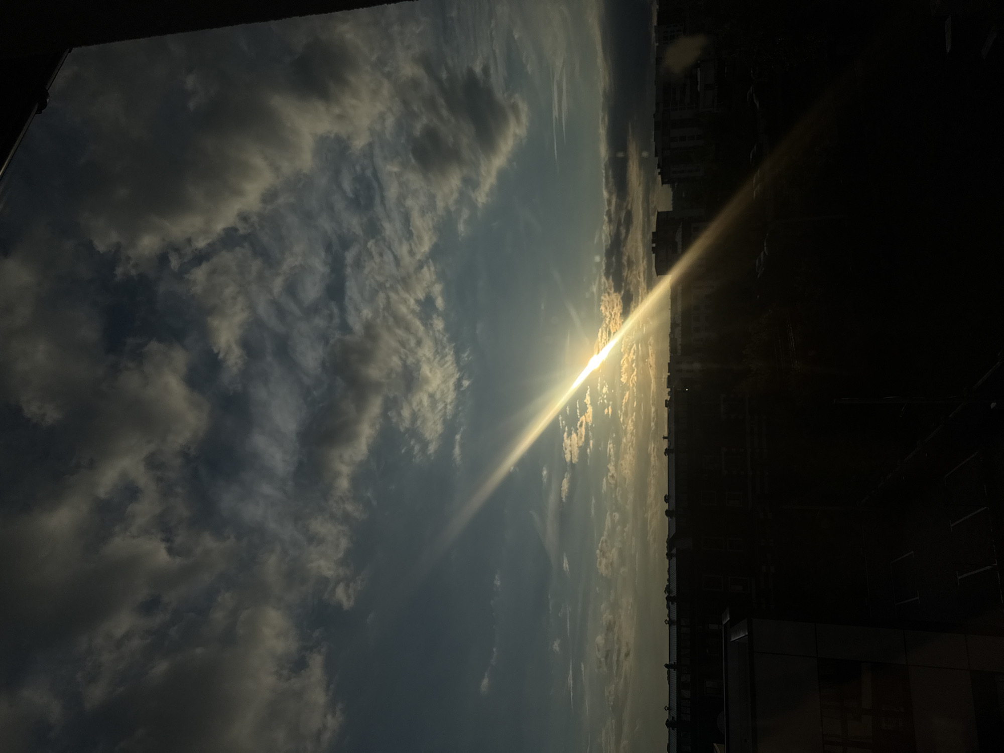 A low-angle shot captures a dramatic sunset over a city. The sun, a brilliant orb, is partially obscured by dark, heavy clouds, casting a sharp, diagonal beam of light across the sky. The light ray illuminates the underside of some of the clouds with a golden hue. The sky transitions from a deep blue to a lighter cyan towards the horizon, where silhouetted buildings form a city skyline. The lower portion of the image is very dark, showing the tops of buildings and an area that appears to be a parking lot, with vehicles vaguely discernible. The scene is framed by a dark, indistinct edge on the right side of the image, possibly a window frame or building structure. The overall mood is somber yet awe-inspiring, emphasizing the power and beauty of natural light.