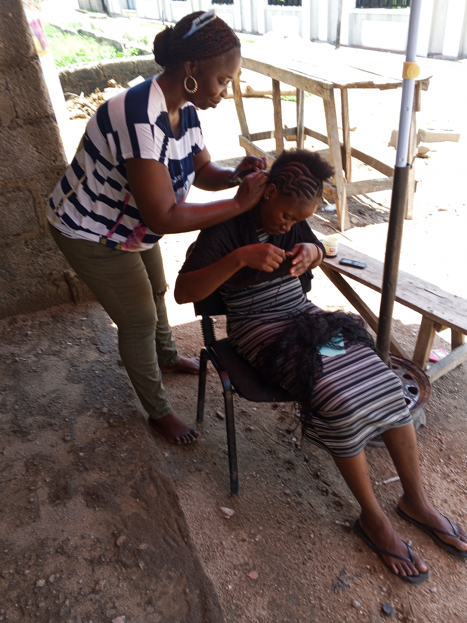 In Zango-ndaji, Nigeria, an outdoor scene depicts a woman standing behind a seated woman, attending to her hair. The standing woman wears a horizontally striped blue and white shirt, olive-green pants, and gold hoop earrings. Her hair is styled in a bun with a headband. She is barefoot. The seated woman is wearing a horizontally striped dress and black flip-flops. She is seated on a black chair. A mass of black hair extensions rests in her lap. A small jar and a cell phone are placed on the wooden bench next to her. The ground is dirt. A wooden table and pole are visible in the background. A concrete structure forms the left-hand edge of the photo.