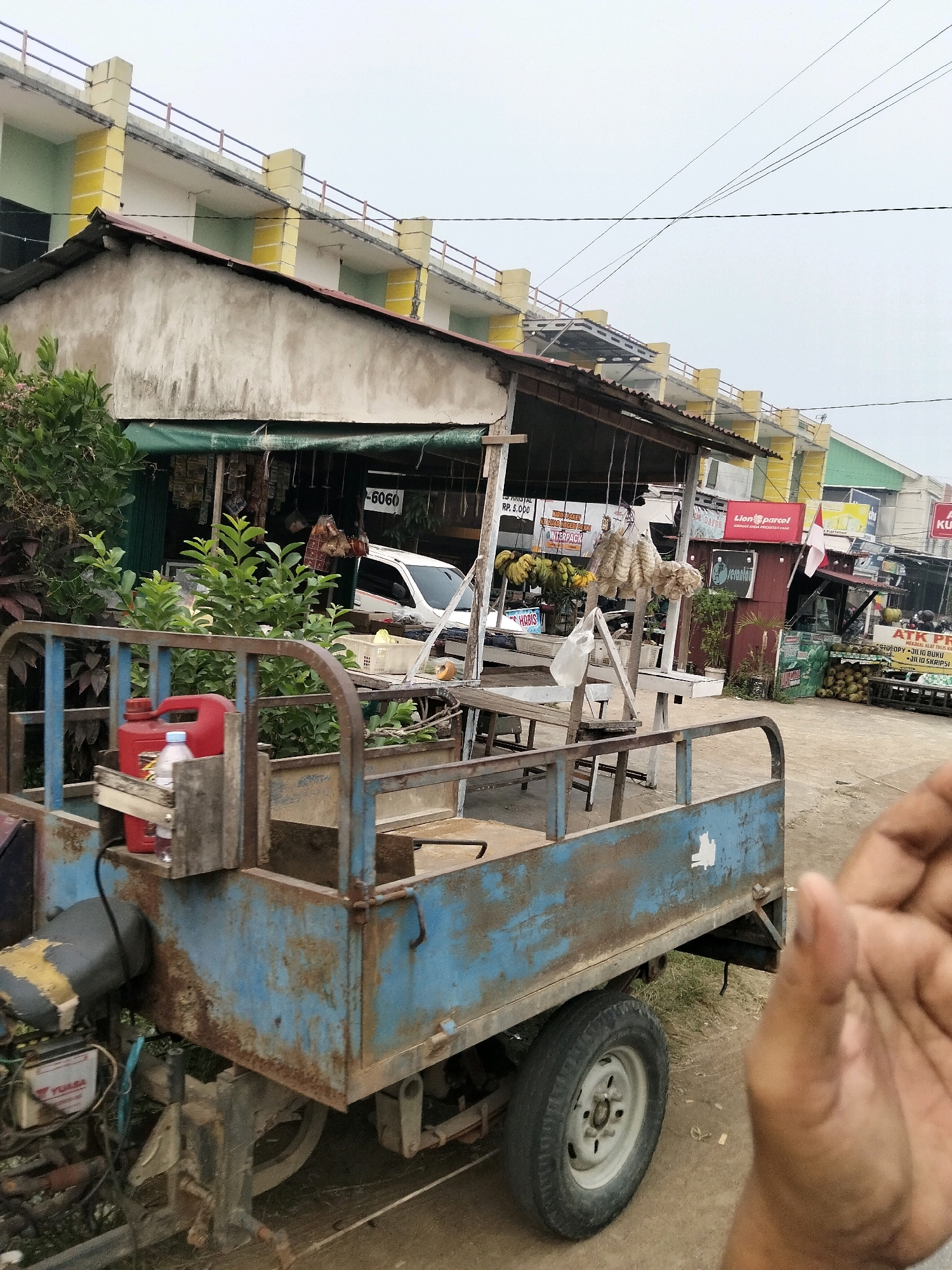 In the city of Arang Limbung, Indonesia, a light blue, weathered motorized cart sits on a dirt road, its engine and rear wheel visible. The cart's bed is empty, except for a red jerrycan and a plastic water bottle. In the background, a market area is set up with stalls. One stall, covered by a green awning, displays various goods, including bunches of bananas and dried items hanging from the ceiling. A white car is parked behind this stall. Another stall features a red sign with a white flag and what appears to be packaged goods. To the right, a large pile of coconuts is stacked. The scene is framed by a multi-story building with a cream-colored facade and yellow pillars, topped with a railing. Power lines are visible crisscrossing the overcast sky. A person's hand is partially visible in the foreground on the right side of the image.