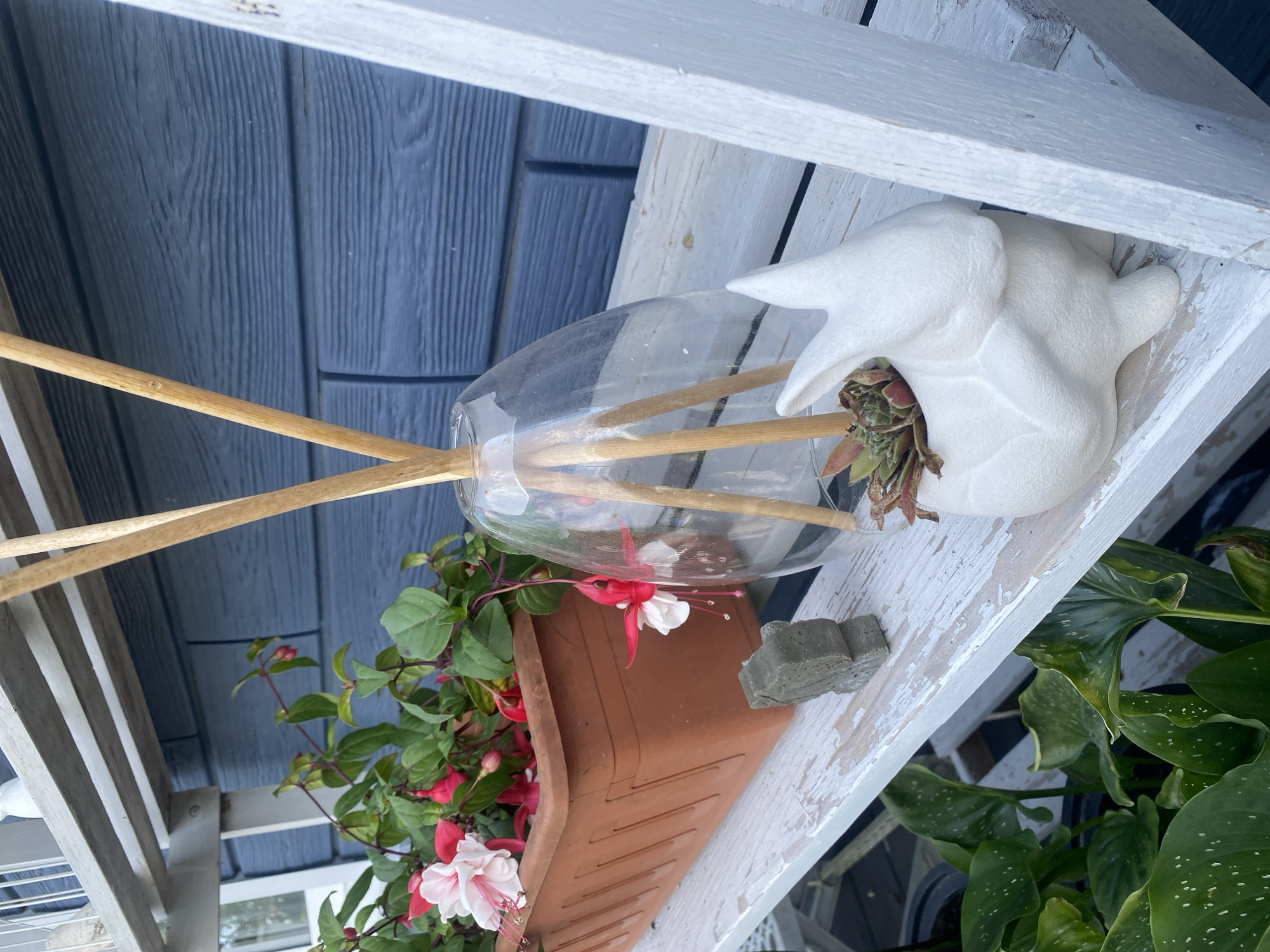 The image depicts a close-up view of a weathered white wooden shelf, possibly part of a tiered plant stand, with items arranged on its surface. The background features a blue, horizontal-slatted wall, characteristic of outdoor siding.
On the shelf, from left to right, are several objects. A rectangular terracotta planter holds flowering Fuchsia plants with pink and white blossoms. Next to the planter is a small, gray, rectangular concrete decorative object. To the right of that is a clear glass vase with several wooden diffuser reeds extending from its opening. Further to the right, a white ceramic planter shaped like a rabbit holds a small succulent plant.
The overall environment suggests an outdoor setting, likely a patio or garden area. The lighting indicates it is daytime. There are no people visible in the image. The location within Tret'ya Pad', Russia, is not discernible from the visual information provided in the image itself.