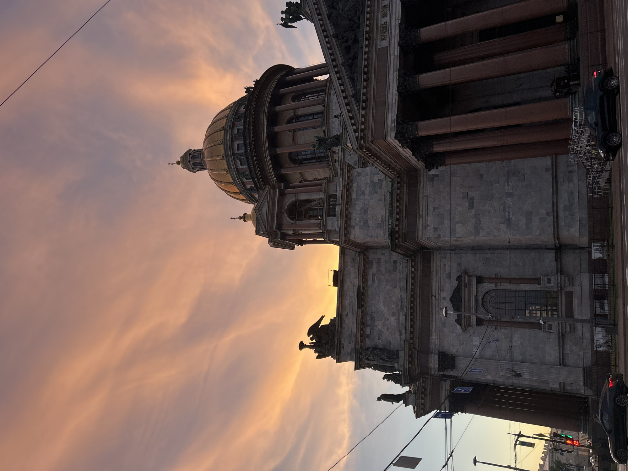 This is a low-angle, wide shot of St. Isaac's Cathedral in Saint Petersburg, Russia, during sunset. The sky is filled with wispy, orange and pink clouds, illuminated by the setting sun. The golden dome of the cathedral is visible in the upper right quadrant, topped with a cross. The facade of the cathedral is made of grey stone and features large columns and several statues. In the foreground, a dark-colored car is parked on the street in front of the cathedral, and another dark-colored car is driving away on the right side of the frame. Traffic lights and utility wires are visible in the background.