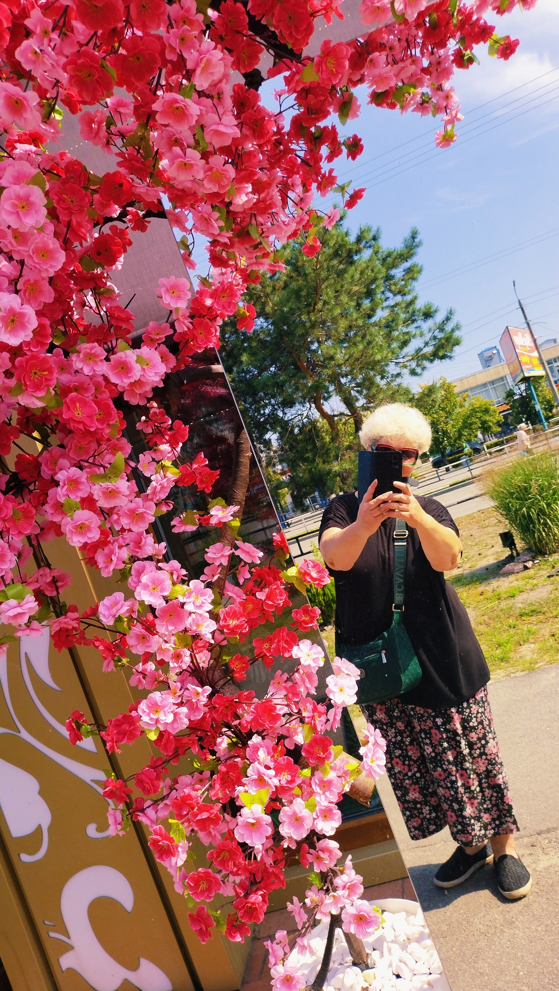 The image captures a woman with a white afro hairstyle and red-rimmed glasses taking a selfie in front of a large mirror adorned with artificial pink and red cherry blossoms. She is wearing a black t-shirt, a floral skirt, and black sneakers, with a green crossbody bag. The mirror reflects a street scene with trees, buildings, and a blue sky, suggesting an outdoor urban environment. The overall setting appears to be in the city of Horishni Plavni, Ukraine, possibly near a commercial area or park, indicated by the presence of buildings and street elements in the reflection. The time of day is likely daytime, given the bright sunlight.