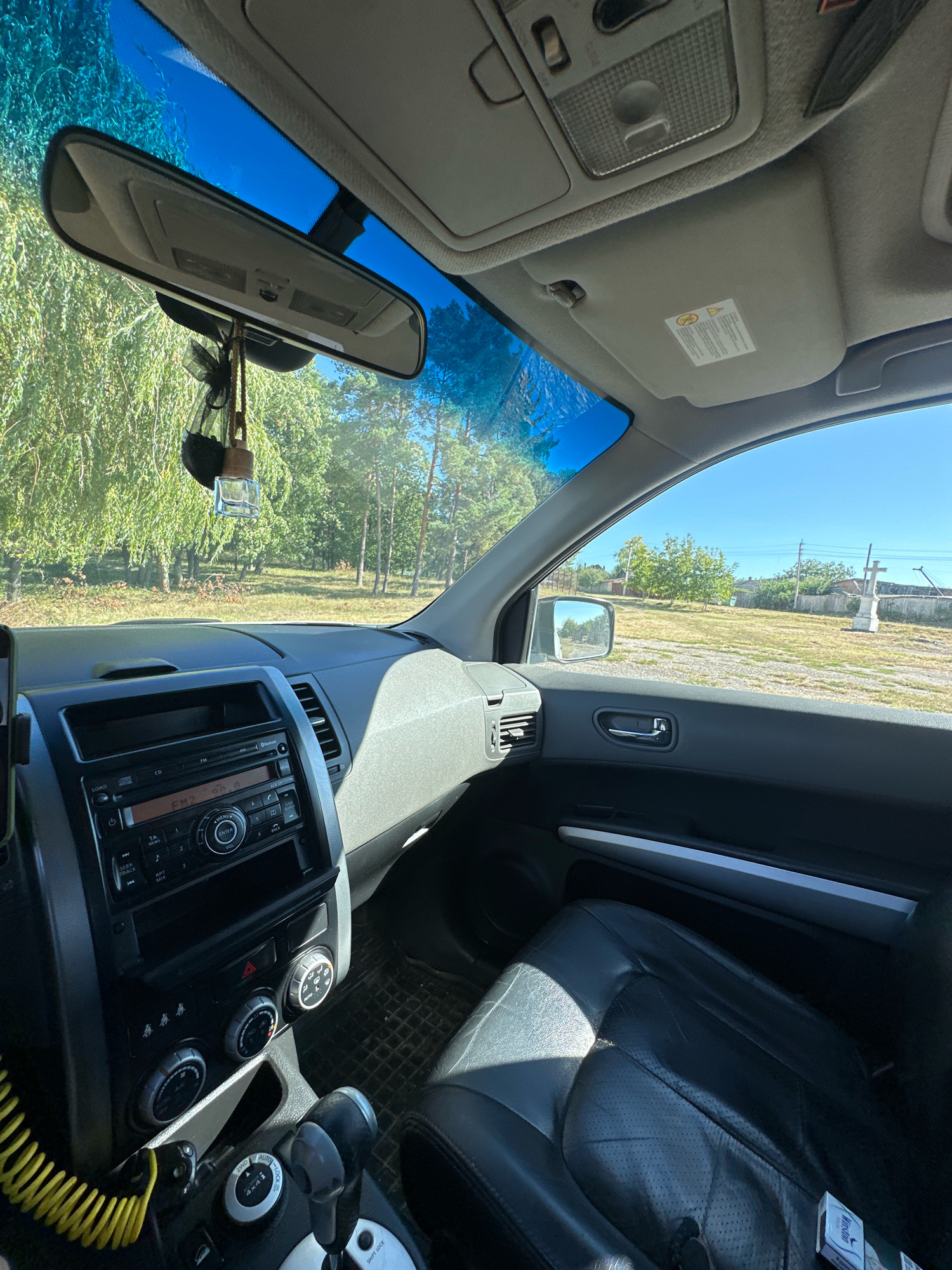 This is a POV shot from inside a car, looking towards the passenger side. The car's interior is visible, including the dashboard with the stereo and climate controls, a leather passenger seat, and the center console with the gear shifter. A car air freshener hangs from the rearview mirror. Outside the car, a rural landscape is visible, with trees, a dirt road, a small building, and a cross. The sky is clear and blue. The location is Makiv, Ukraine.