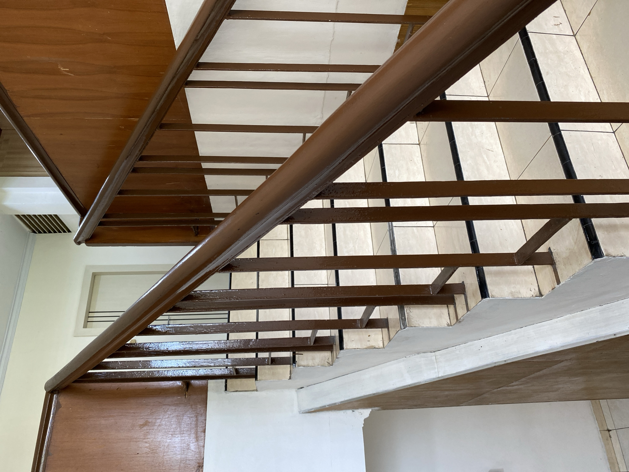 The image shows an interior view of a staircase with wooden handrails and railings. The steps are tiled with light-colored tiles and bordered with black trim. The walls are painted white, and there is wooden paneling along the staircase. In the background, a doorway with vertical blinds is visible. The scene appears to be an indoor public space, possibly an office building or residential complex, located in Banda Aceh, Indonesia. There are no people or actions depicted in the image.