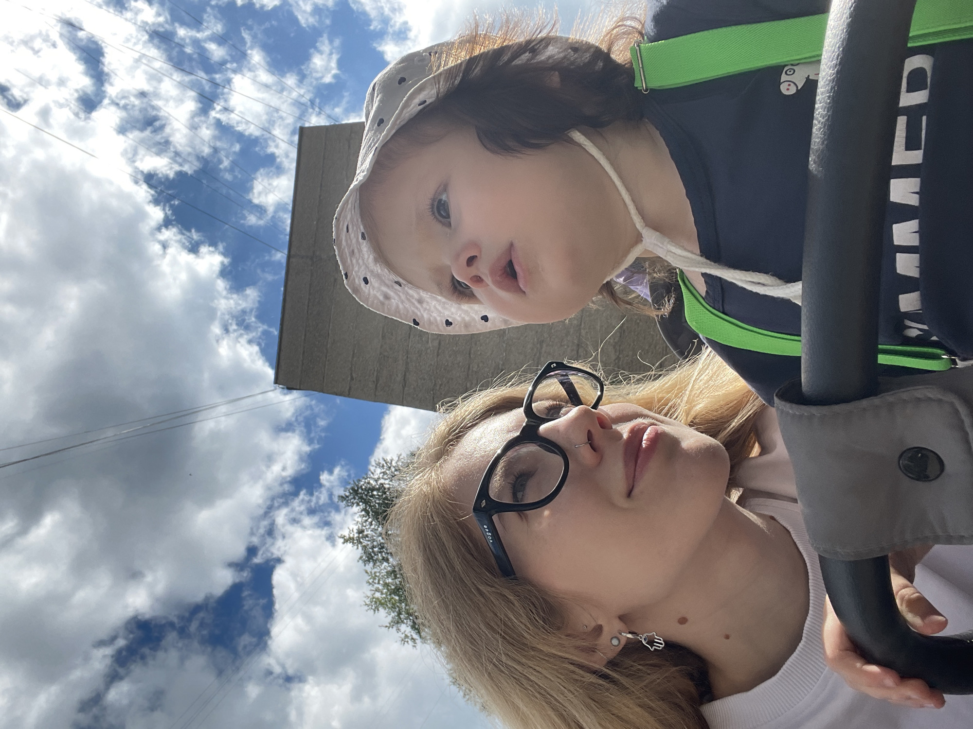 The image shows a close-up of a young woman and a toddler in what appears to be an outdoor urban setting in Izhevsk, Russia. The woman, with blonde hair, is wearing glasses and a white sweatshirt, and has a hamsa hand earring. She is looking to the side with a slight smile. The toddler, wearing a white hat with black polka dots and a dark blue t-shirt with green suspenders, is looking upwards with their mouth slightly open. They are seated in a stroller, with the stroller's handle visible in the foreground. In the background, there is a concrete structure and a bright blue sky with clouds and overhead power lines, suggesting the location could be near industrial or infrastructural areas in the city. The lighting indicates it is daytime, likely a sunny day given the bright sky and shadows.