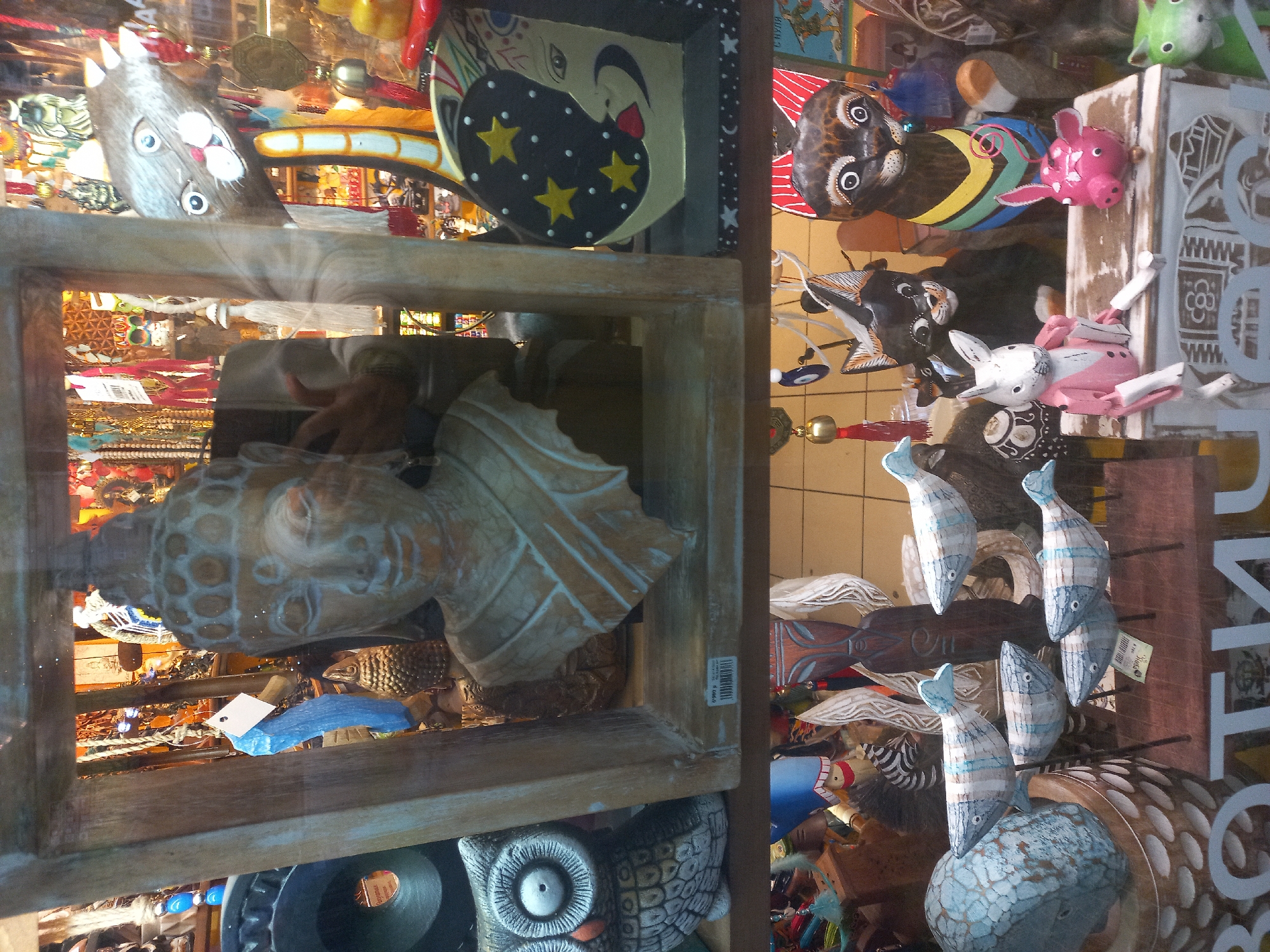 A shop window in Sochi, Russia, is filled with various handcrafted items. On the left, a wooden frame displays a Buddha head and a leaf sculpture, with a cat head above and an owl below. Other items include a crescent moon face and a variety of animal figurines such as cats, pigs, and rabbits. Wooden fish are displayed on the right side of the window.