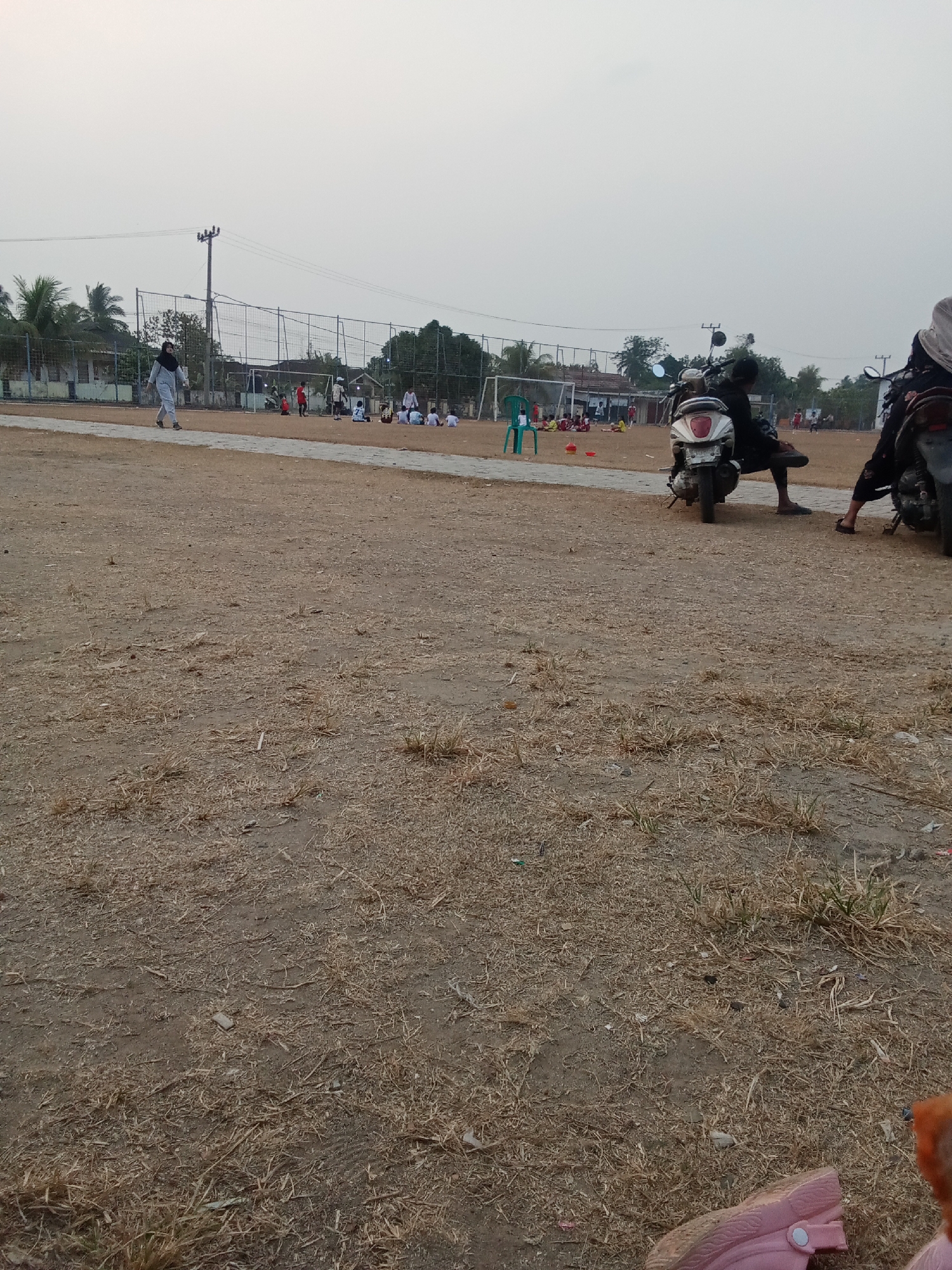 In Toto Katon, Indonesia, an outdoor scene unfolds on a dry, dirt field, possibly a sports ground, under an overcast sky. A chain-link fence and a goalpost suggest a soccer field. In the middle ground, several people are gathered, some appearing to be children sitting on the ground, while others stand and observe. A woman in a hijab walks across the field. Two motorcycles are parked in the foreground, one with a person seated on it. The ground is covered with dry grass and scattered debris.